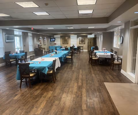 A spacious dining room with several tables covered in blue and white tablecloths, each surrounded by wooden chairs. The room has wooden flooring, beige walls, and multiple windows letting in natural light. There are framed pictures on the walls and a clock on the right side wall.