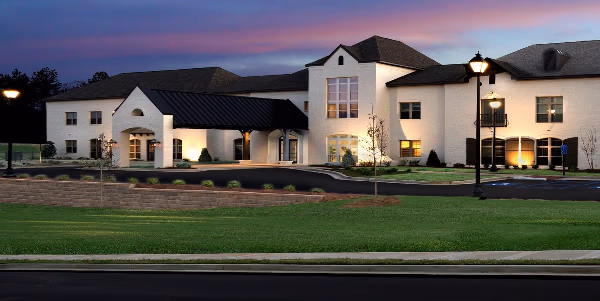 Two-story white brick senior living building at dusk with a covered entrance, illuminated windows, lampposts, driveway, and manicured lawn.