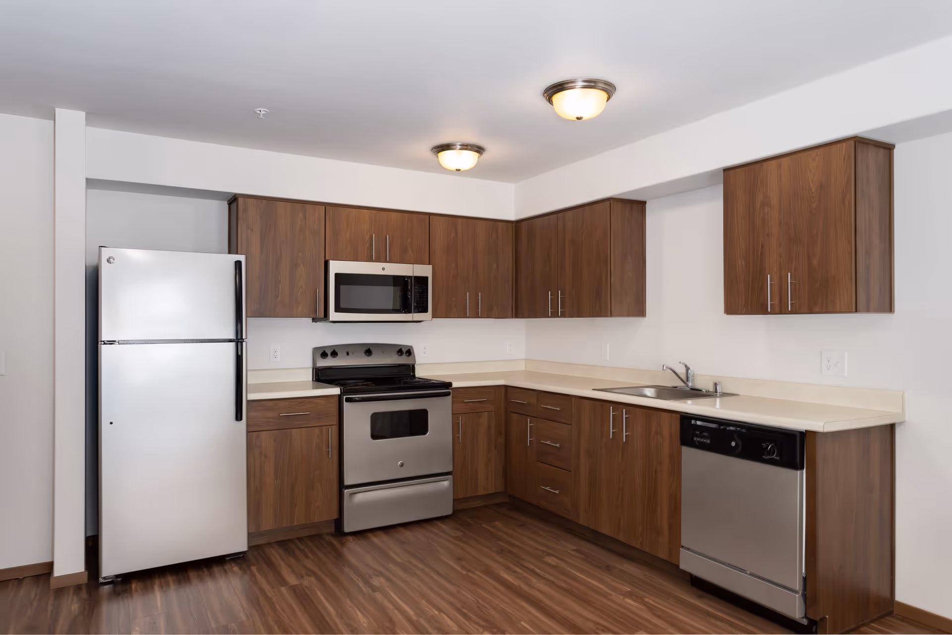 A modern kitchen with wooden cabinets, stainless steel appliances including a refrigerator, microwave, oven, and dishwasher, and a double sink. The kitchen has a light-colored countertop and wood flooring.