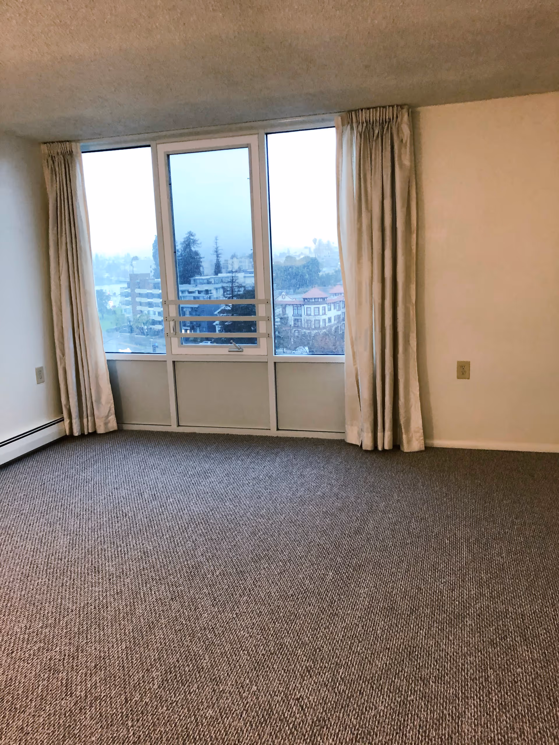 Empty room with beige carpet and cream-colored walls featuring a large window with three glass panels and beige curtains on either side, overlooking a cityscape on a cloudy day.