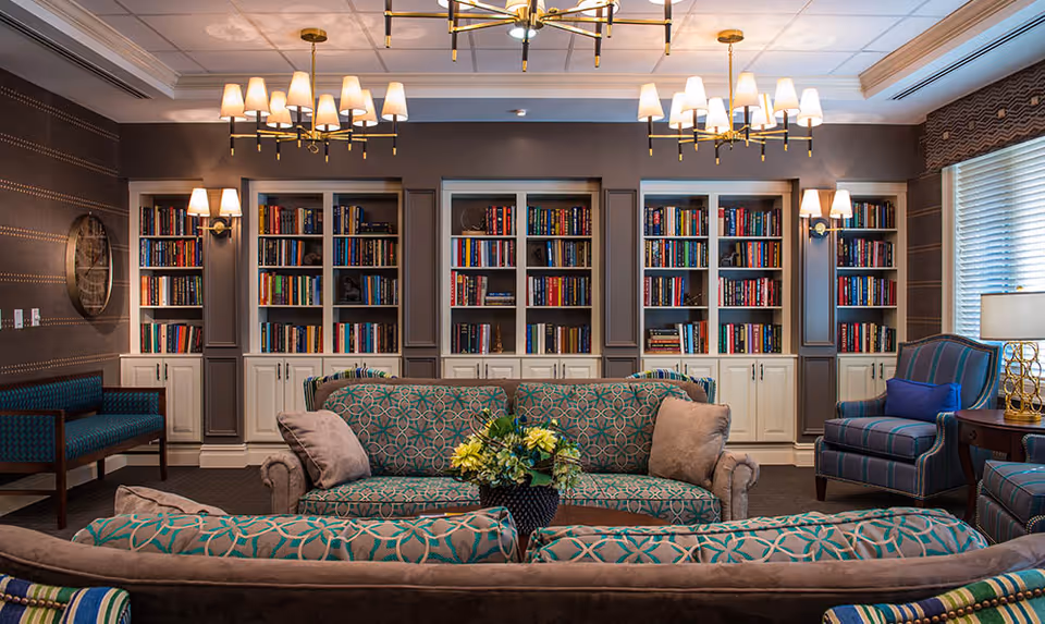 A cozy and elegant living room area with patterned sofas and armchairs arranged around a central coffee table with a flower arrangement. Behind the seating area is a wall with built-in bookshelves filled with books, flanked by wall sconces and decorative lighting fixtures on the ceiling. Large windows with blinds allow natural light into the room.
