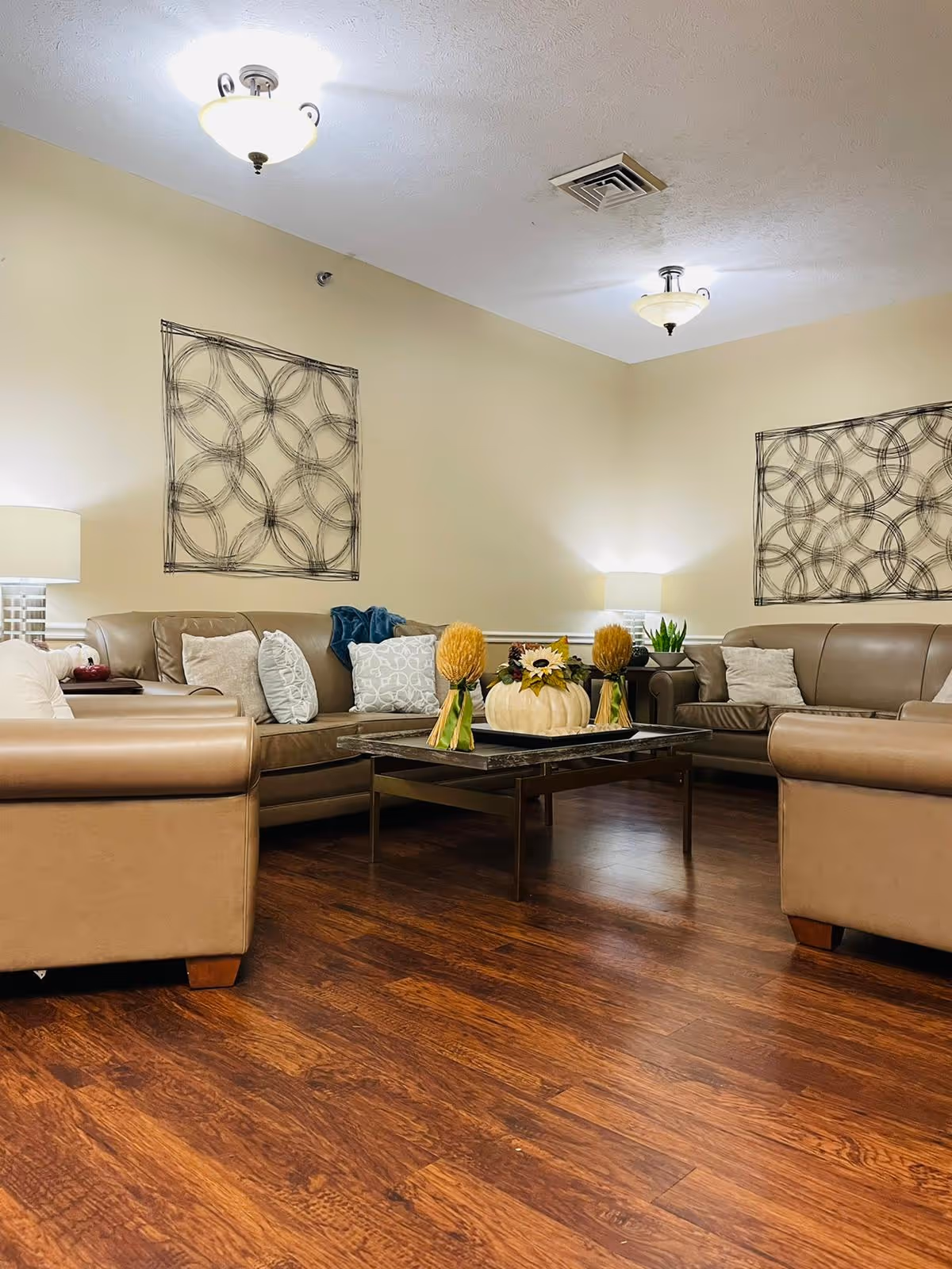 A cozy living room with brown leather sofas arranged around a rectangular coffee table. The coffee table has decorative items including a white pumpkin and yellow floral arrangements. The room features wooden flooring, beige walls, two table lamps on side tables, and abstract circular wall art.