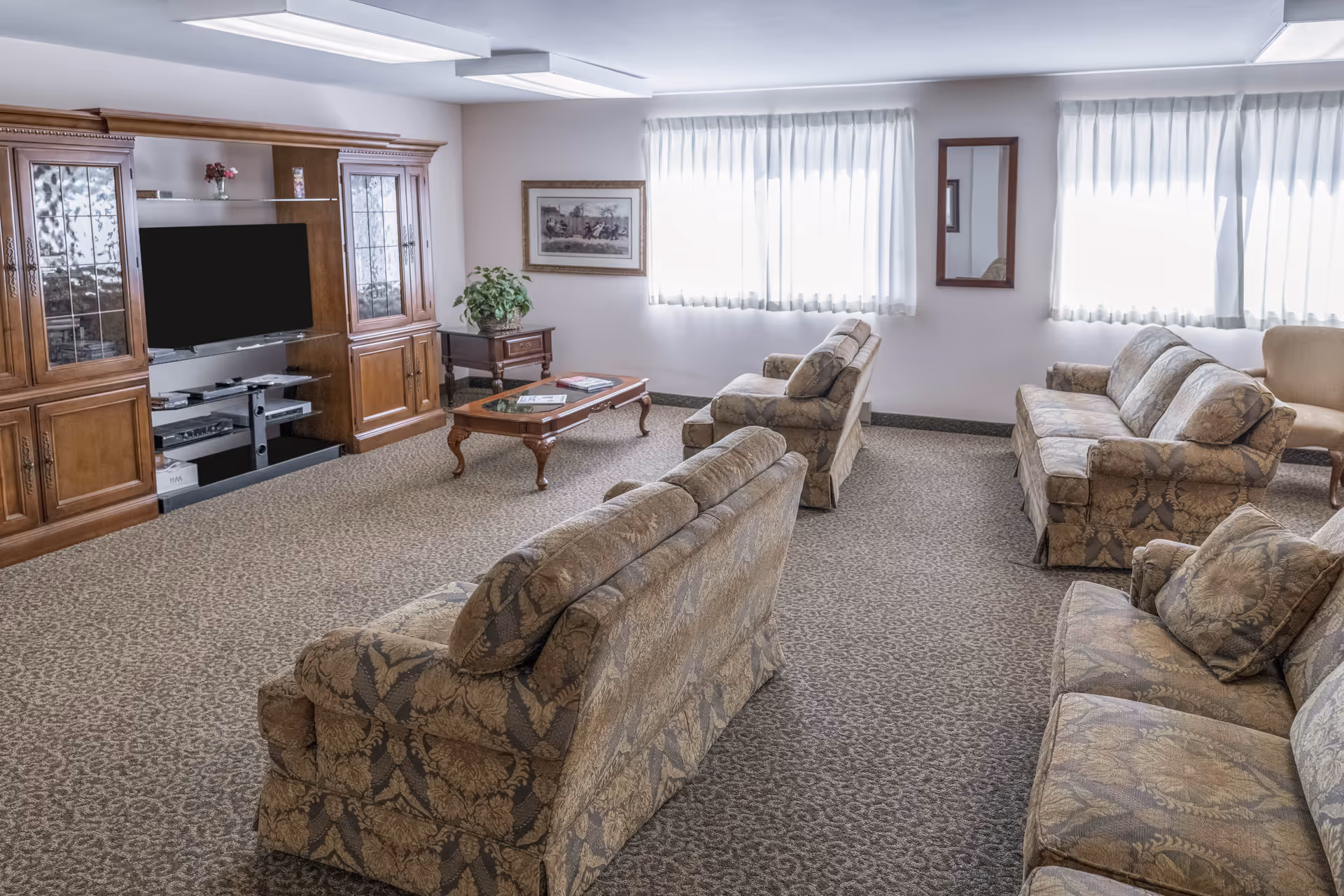A spacious living room with patterned upholstered sofas and armchairs arranged around a wooden coffee table. The room features a large wooden entertainment center with a flat-screen TV, a side table with a plant, framed artwork on the wall, and two large windows with white curtains letting in natural light.