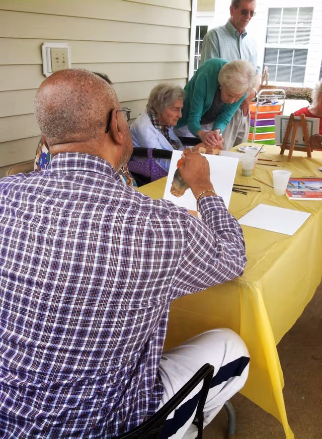 A group of elderly people sitting around a table covered with a yellow tablecloth, engaging in an art activity. One man in a plaid shirt is painting on a piece of paper, while others watch and interact. The setting appears to be an outdoor covered patio area with beige siding on the wall.