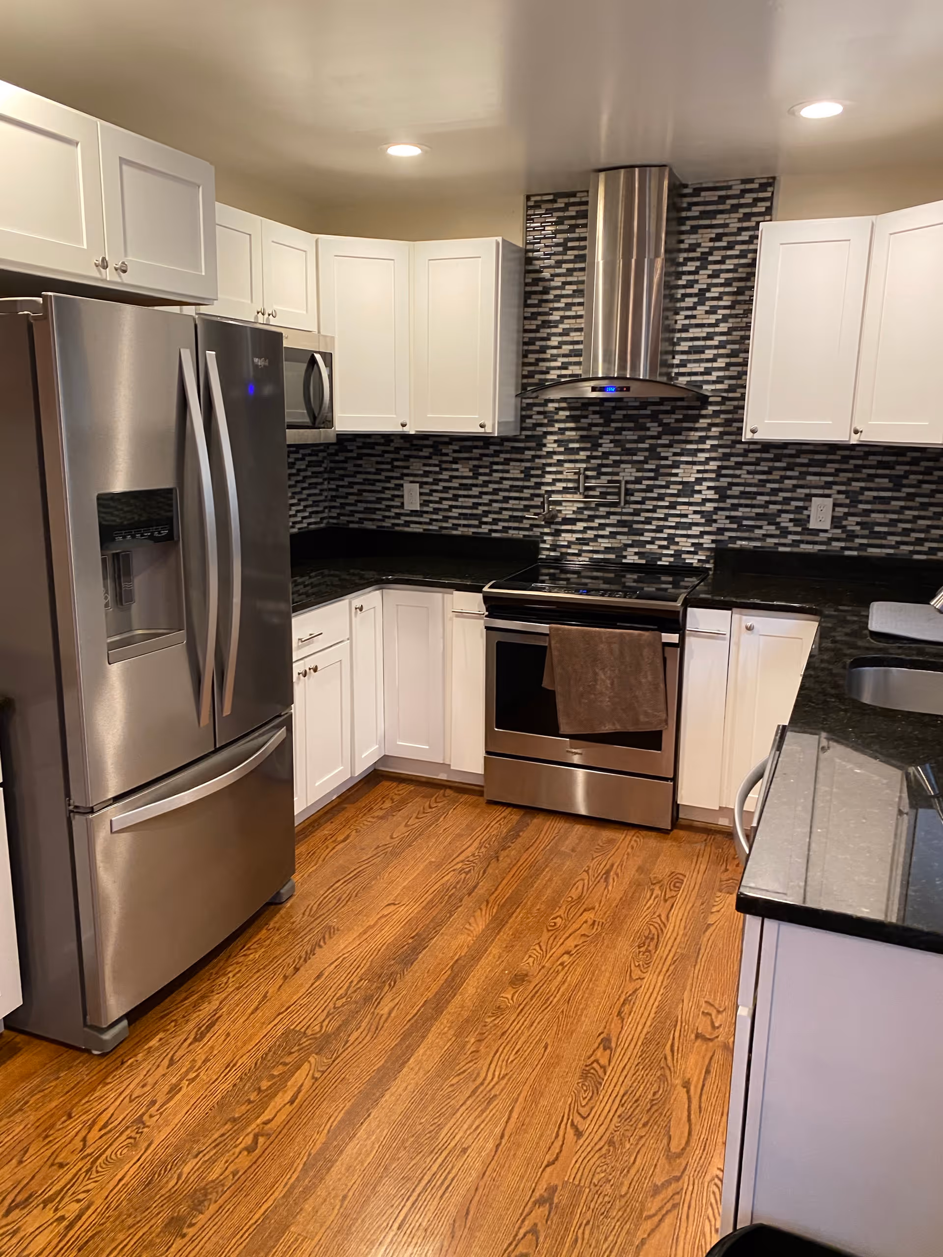 Modern kitchen with stainless steel refrigerator, oven, and microwave. White cabinets with silver handles, black granite countertops, and a mosaic tile backsplash in shades of black, white, and gray. Hardwood flooring and a stainless steel range hood above the stove.
