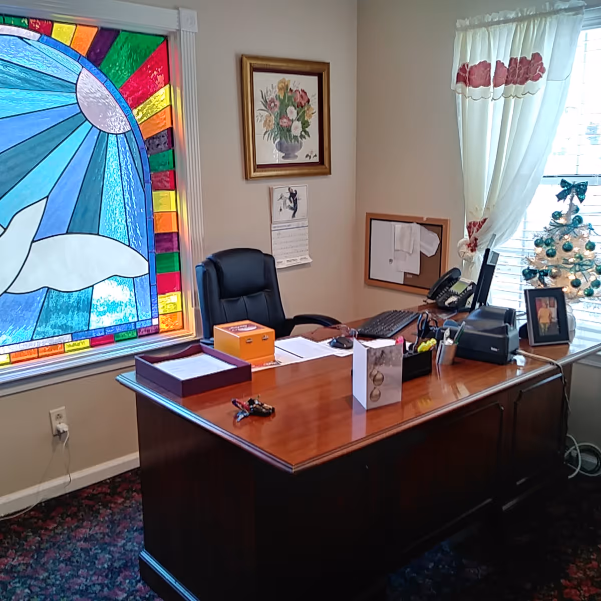 Office interior with a wooden desk, black chair, stained glass window, and a small decorated Christmas tree by the window.
