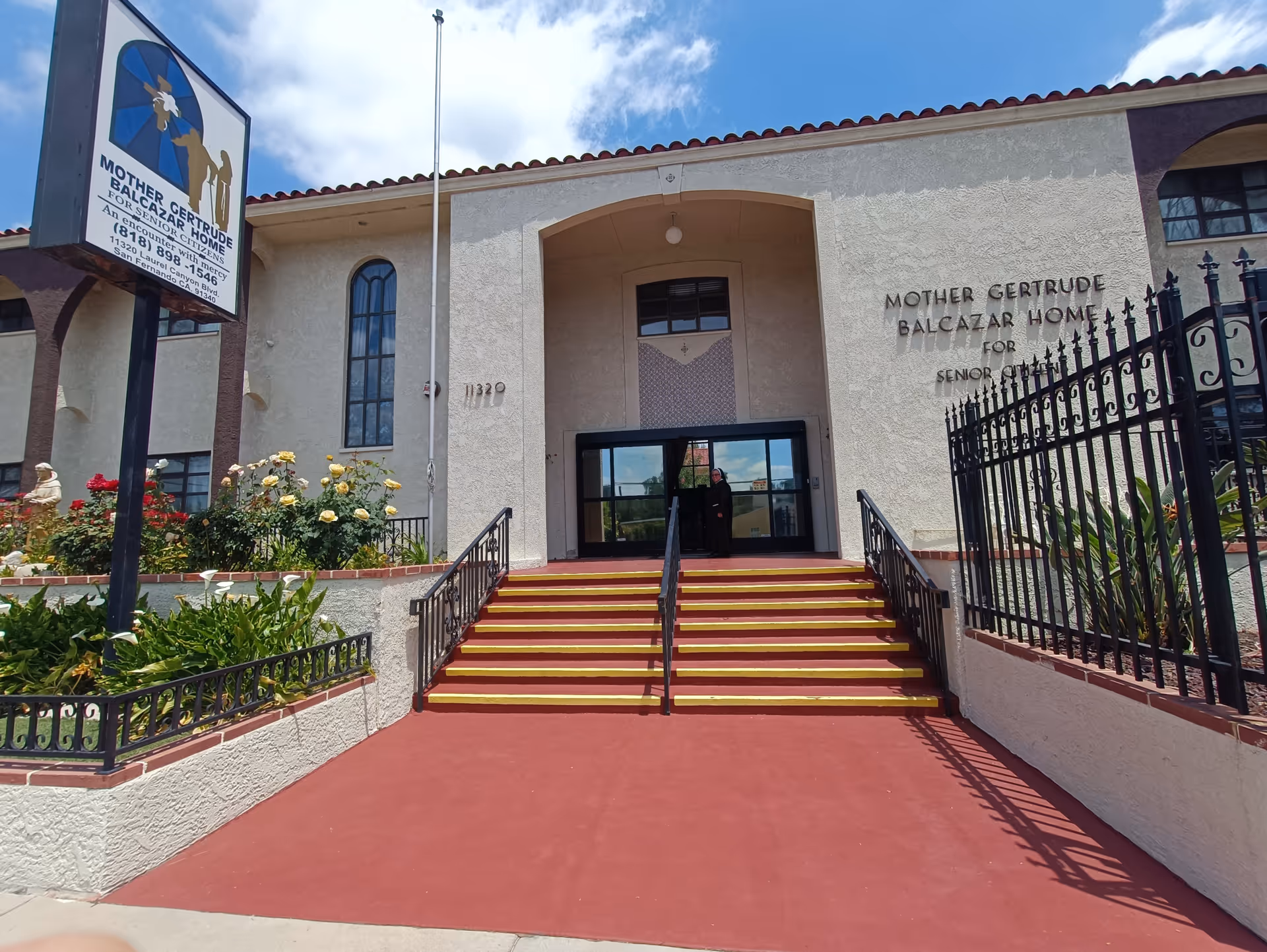 Front entrance of Mother Gertrude Balcazar Home for Senior Citizens, showing a beige building with a red-tiled roof, stairs with yellow safety strips leading up to glass doors, a sign with the facility's name and contact information, and a garden with flowers and greenery on the left side.