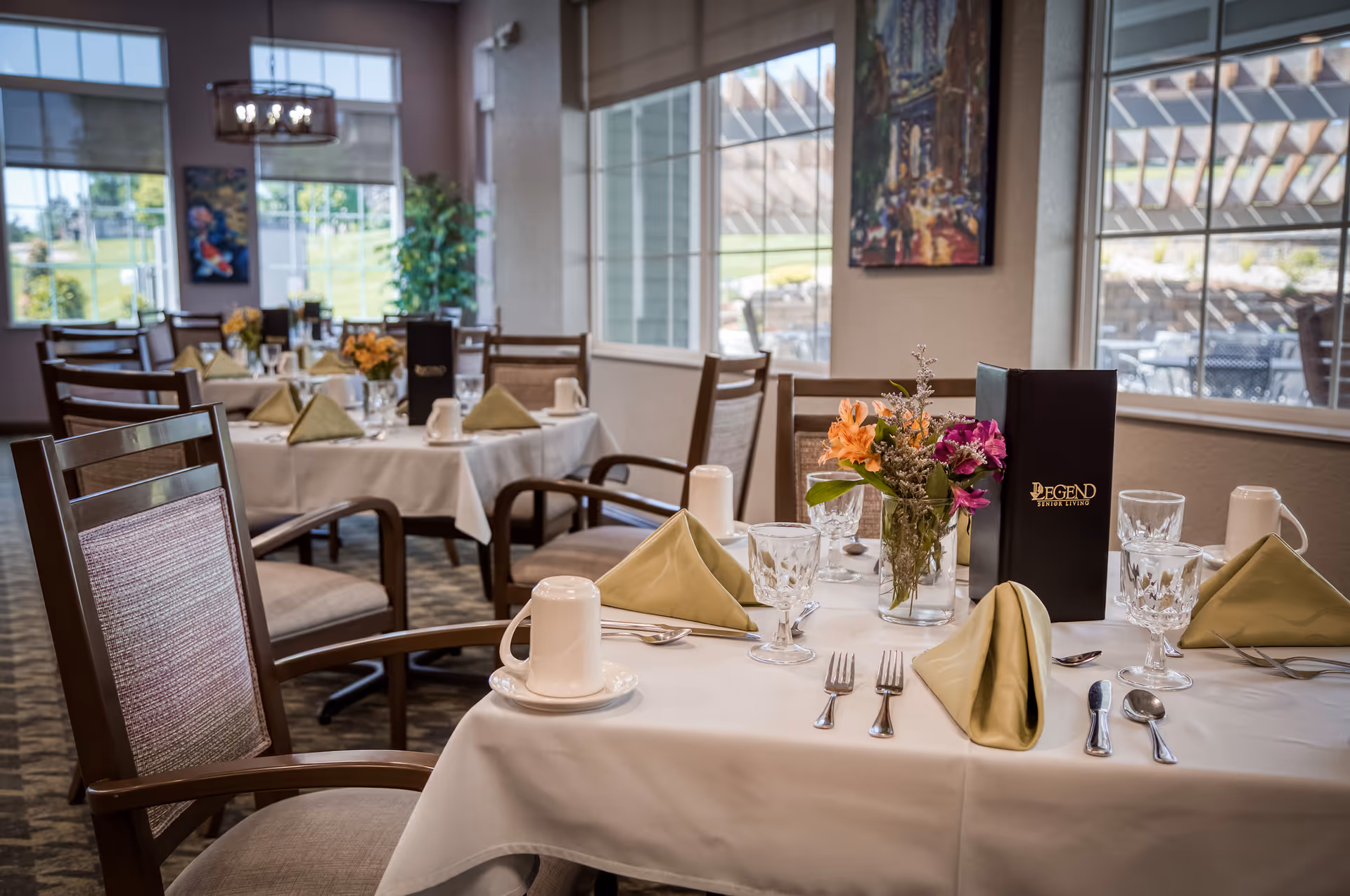 A dining room with tables set for a meal, featuring white tablecloths, folded green napkins, glassware, silverware, coffee cups, and floral centerpieces. Large windows allow natural light to fill the room, and wooden chairs with cushioned seats surround the tables. A menu labeled 'Legend Senior Living' is visible on one table.