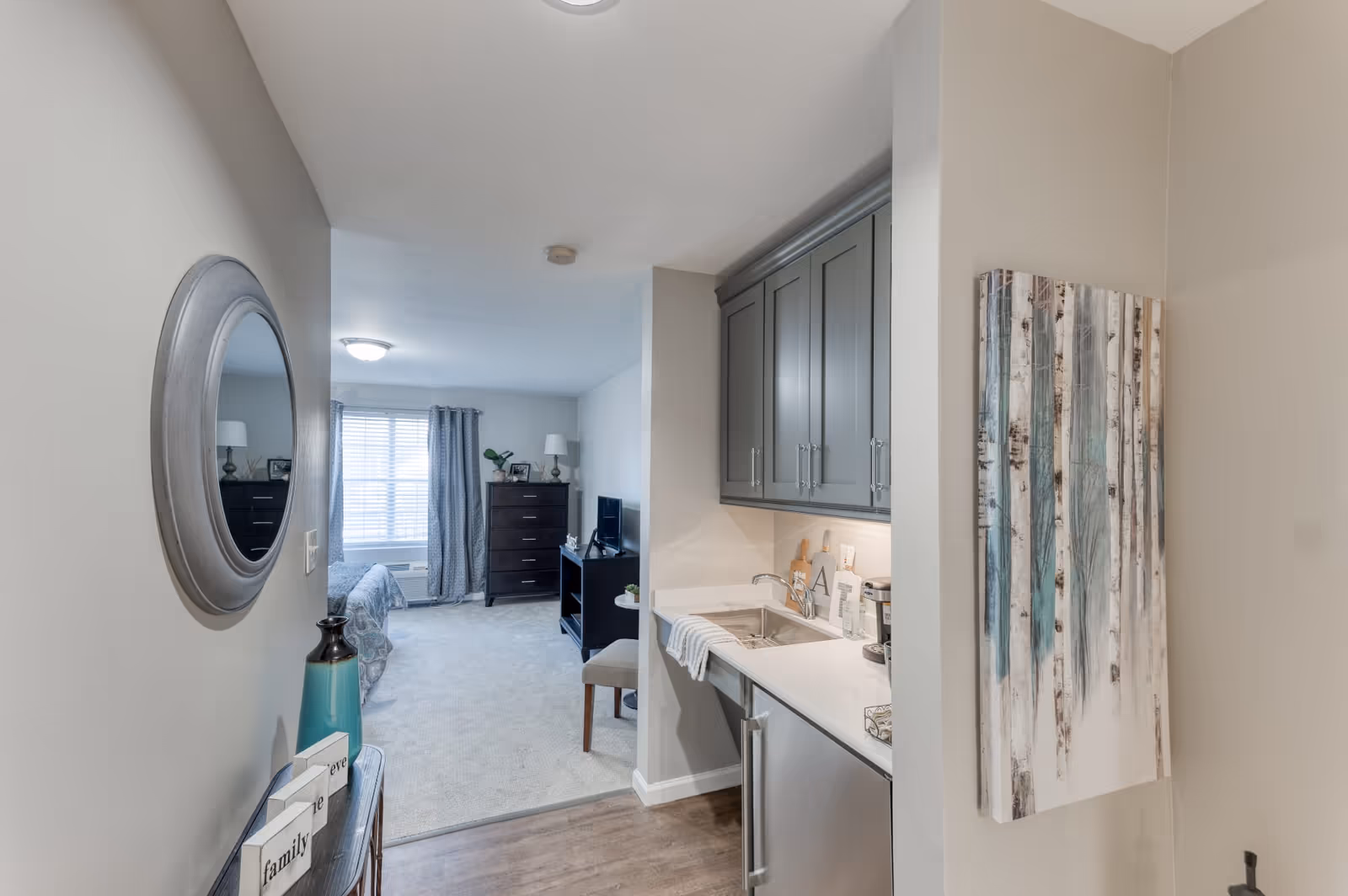 View of a senior living facility room showing a small kitchenette with gray cabinets, a sink, and a mini refrigerator on the right. The room extends into a living and sleeping area with a bed, dresser, TV on a stand, and a window with curtains. A round mirror and decorative items are on the left wall, and a painting of trees hangs on the right wall.