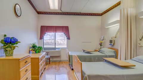 A bright and clean hospital-style room with two beds, each equipped with a wooden overbed table. The room has light-colored walls with a decorative border near the ceiling, a window with a red valance, and wooden dressers with potted plants on top. A clock is mounted on the wall, and there is a privacy curtain partially visible on the right side.