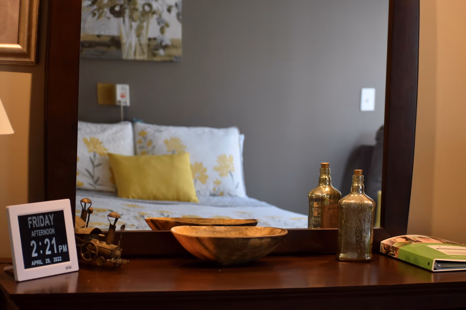 A wooden dresser with a large mirror reflecting a bed with white and yellow floral bedding and a yellow pillow. On the dresser are decorative items including a digital clock displaying the date and time, a small metal train figurine, a wooden bowl, two glass bottles with cork stoppers, and a green binder.