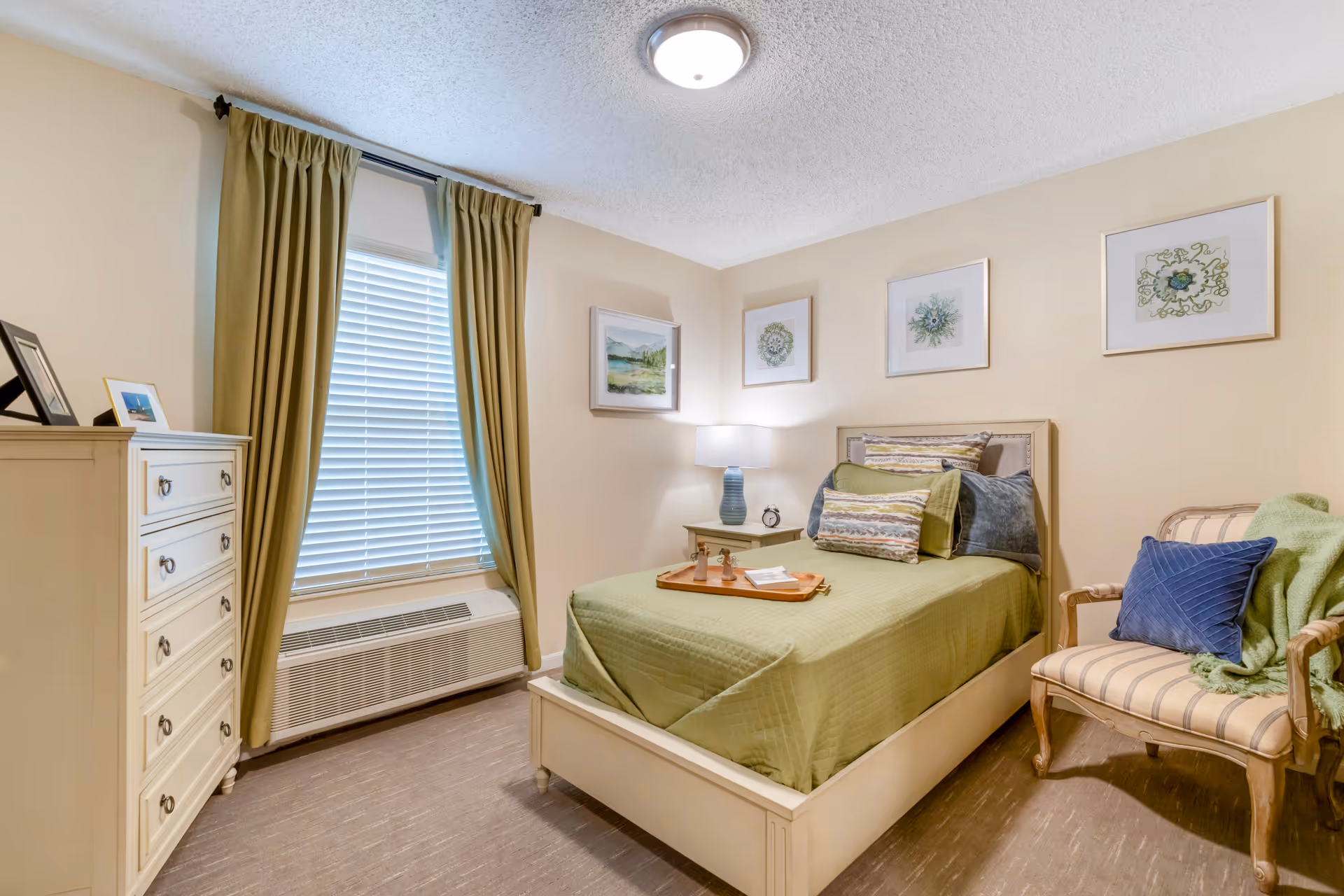 Neat bedroom with a twin bed dressed in green bedding, a nightstand and lamp, dresser, window with green curtains, and an upholstered chair.