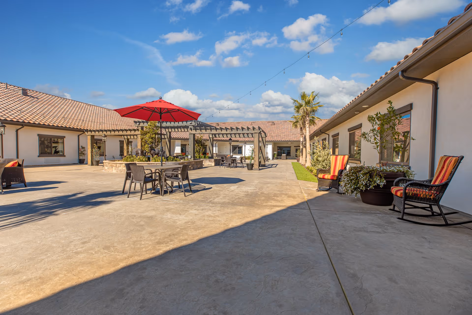 Outdoor courtyard area of a senior living facility with concrete flooring, patio tables and chairs, a red umbrella, potted plants, and cushioned rocking chairs. The area is surrounded by single-story buildings with tiled roofs under a blue sky with scattered clouds.