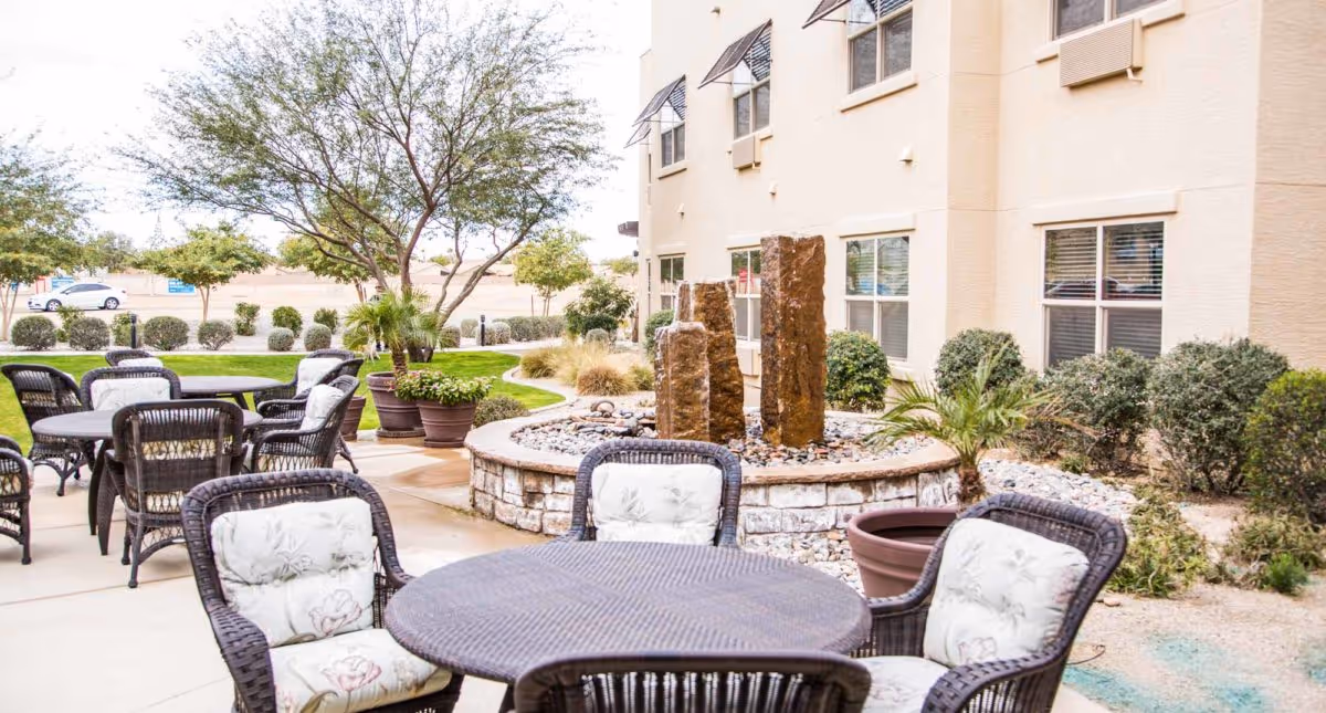 Outdoor patio area with round tables and cushioned wicker chairs arranged on a concrete surface. In the background, there is a stone water feature with three vertical stone pillars surrounded by small rocks. The patio is adjacent to a beige building with multiple windows, and there are various potted plants, shrubs, and trees around the area.