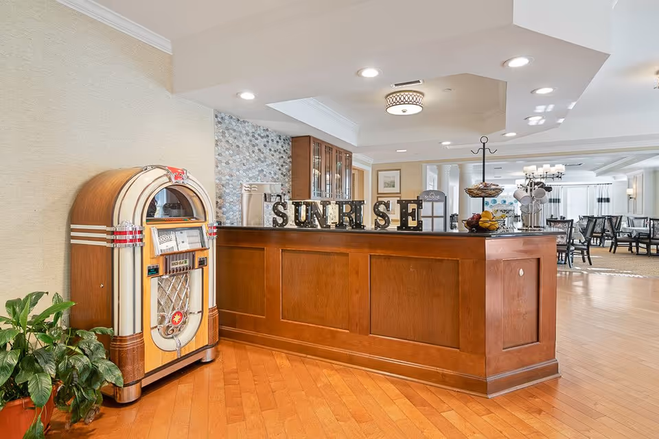 Interior view of a senior living facility reception area with a wooden counter displaying the word 'SUNRISE' in large decorative letters. To the left of the counter is a vintage jukebox and a green potted plant. The background shows a dining area with tables and chairs, bright lighting, and a clean, welcoming atmosphere.