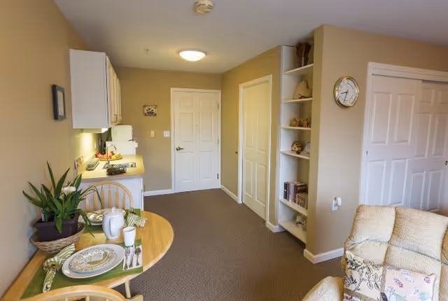 Interior view of a cozy assisted living apartment featuring a small dining table set for two with plates, cups, and a plant. The room includes a kitchenette with white cabinets, a microwave, and a refrigerator. There is a beige carpeted floor, built-in shelves with decorative items and books, a wall clock, and a comfortable armchair partially visible on the right.