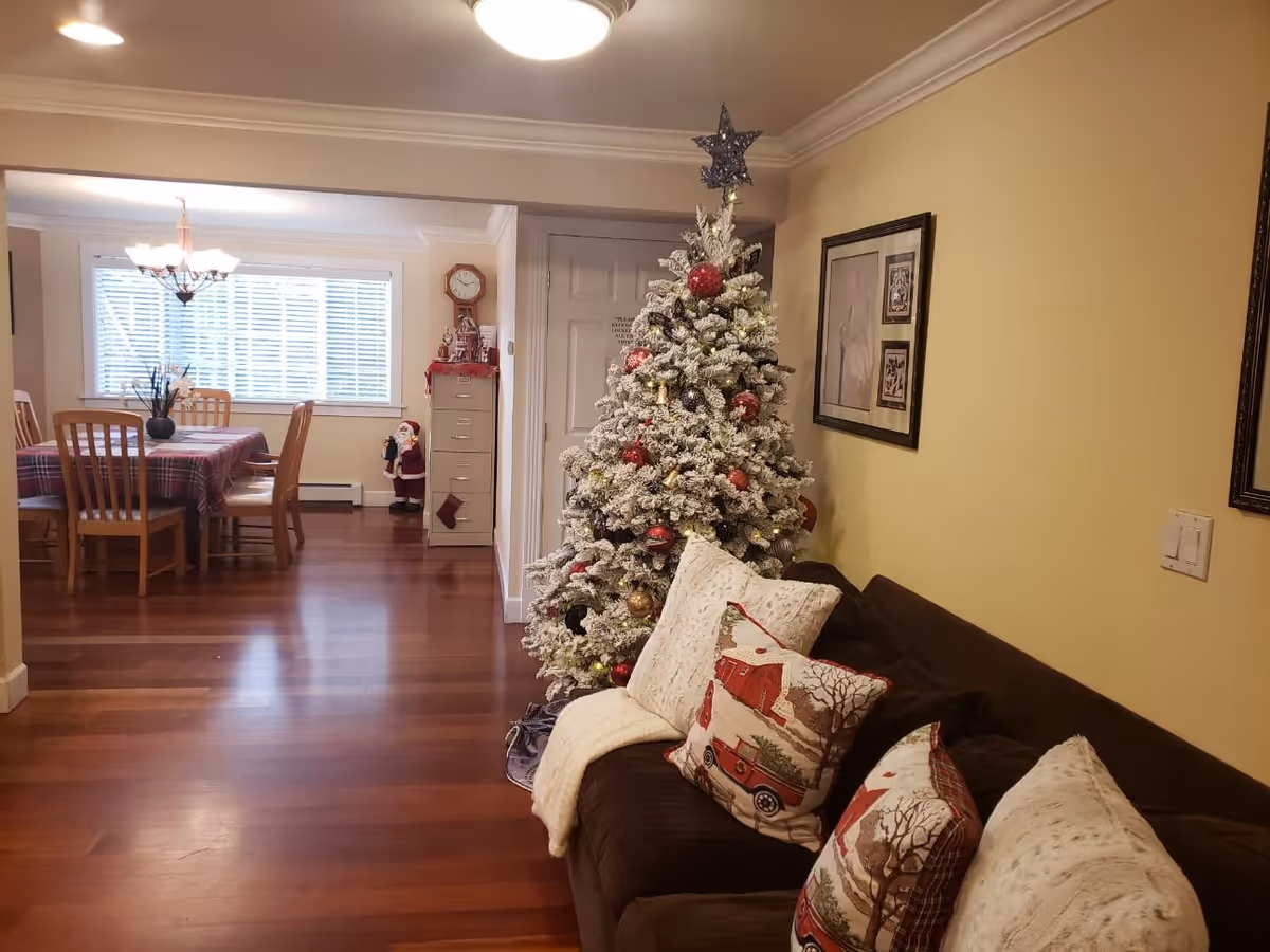 A cozy living room and dining area decorated for Christmas. The living room features a brown couch with festive pillows and a white blanket, next to a decorated Christmas tree with red and gold ornaments and a star on top. The dining area has a wooden table covered with a plaid tablecloth and surrounded by wooden chairs. A chandelier hangs above the dining table, and a window with white blinds lets in natural light. There is a clock and a Santa Claus decoration near a filing cabinet in the background.