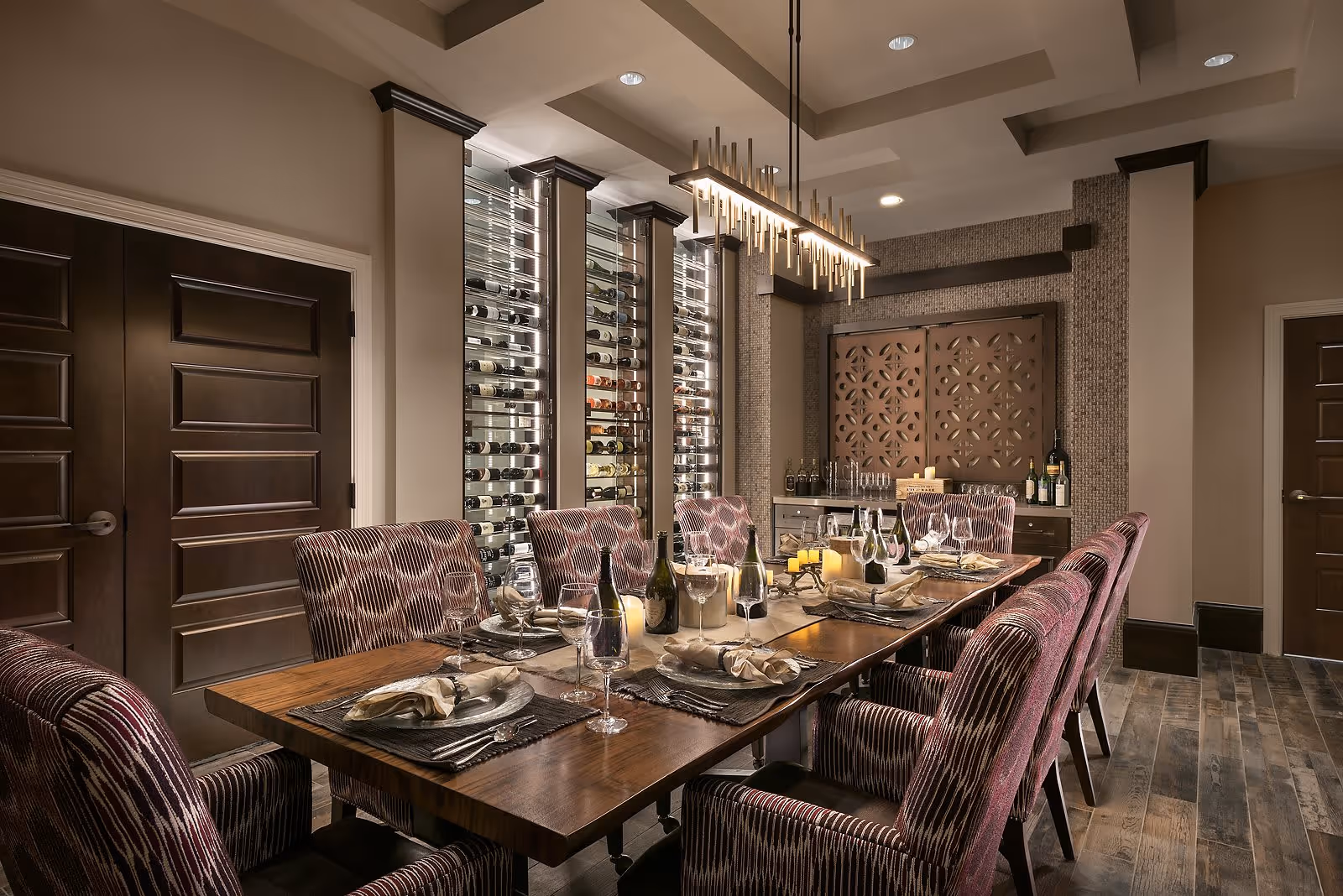 Elegant dining room with a long wooden table set for a meal, featuring wine bottles, glasses, and candles. The room has patterned upholstered chairs, a modern chandelier, wine racks on the wall, and decorative wooden panels. The floor is wooden, and the walls are painted in neutral tones.