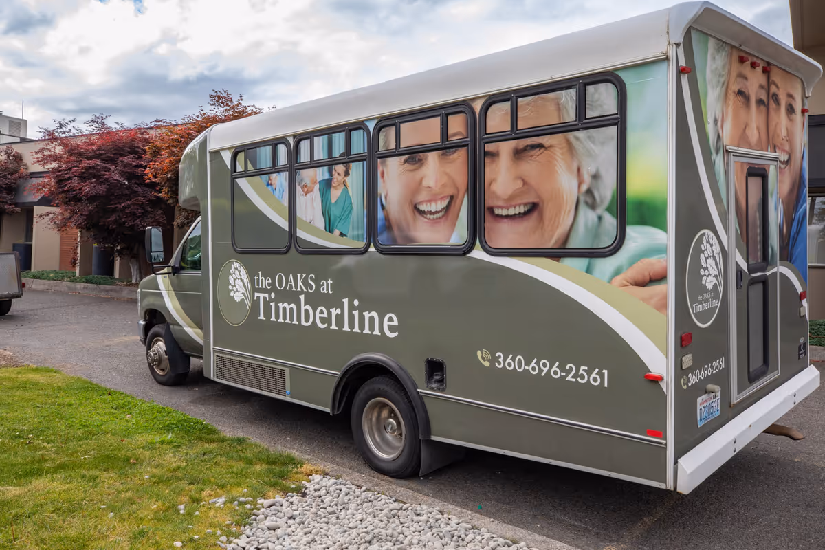 A shuttle bus parked outside a building with images of smiling elderly people and caregivers on its side. The bus is branded with the logo and name 'the OAKS at Timberline' along with a phone number 360-696-2561. The surrounding area includes a small patch of grass, a rock border, and some trees with red leaves.