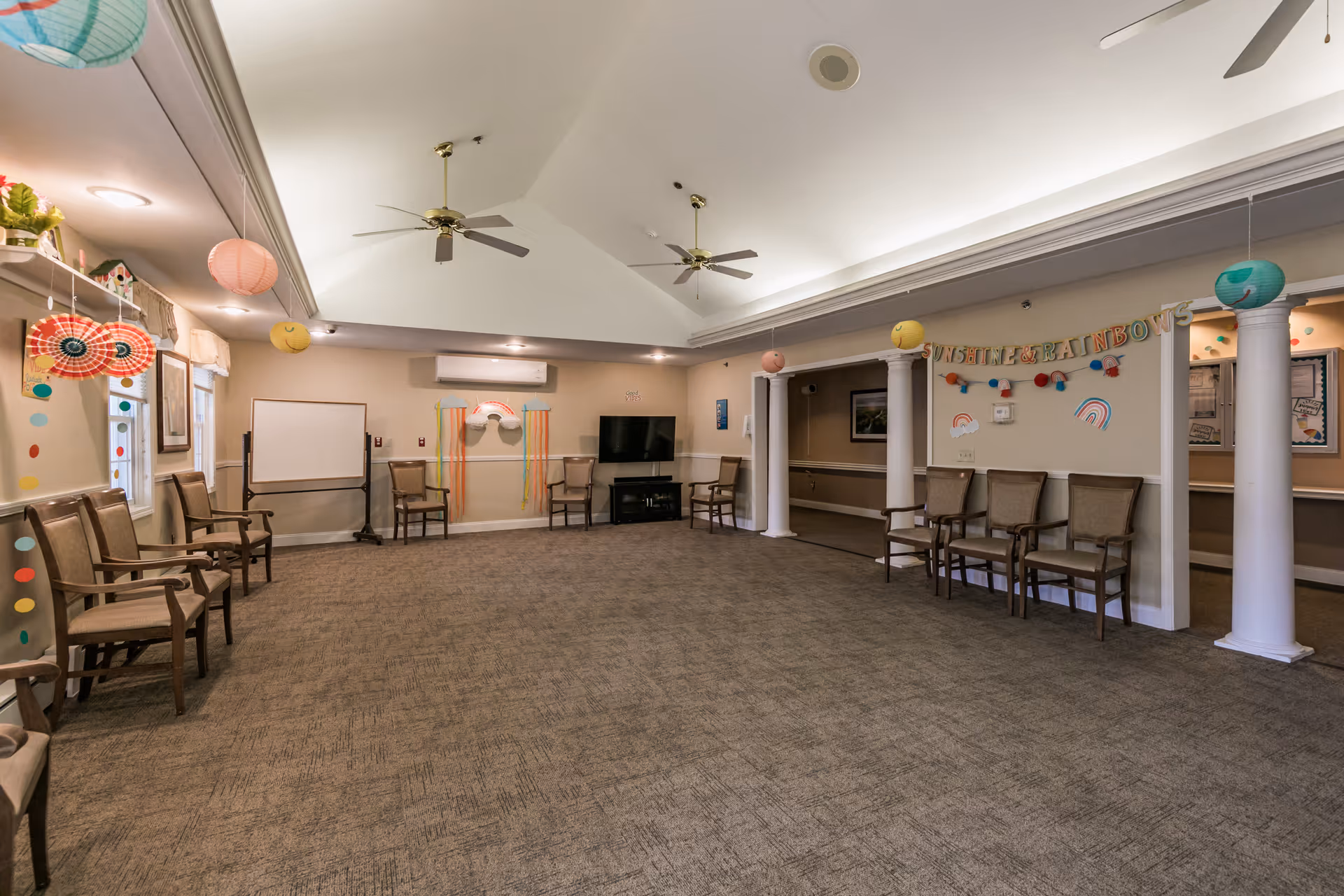 A spacious room with beige walls and carpeted floor, arranged with chairs along the walls. The ceiling is vaulted with two ceiling fans. There are colorful paper lanterns and decorations hanging from the ceiling and walls, including a banner that reads 'SUNSHINE & RAINBOWS'. A flat-screen TV is mounted on one wall above a black cabinet, and a whiteboard is positioned on a stand. The room has a welcoming and communal atmosphere.