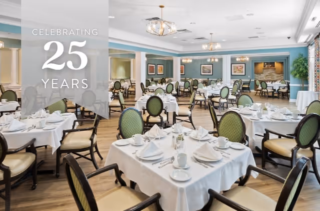 Bright dining room with round tables set with white linens and green-backed chairs and a banner reading "Celebrating 25 Years".