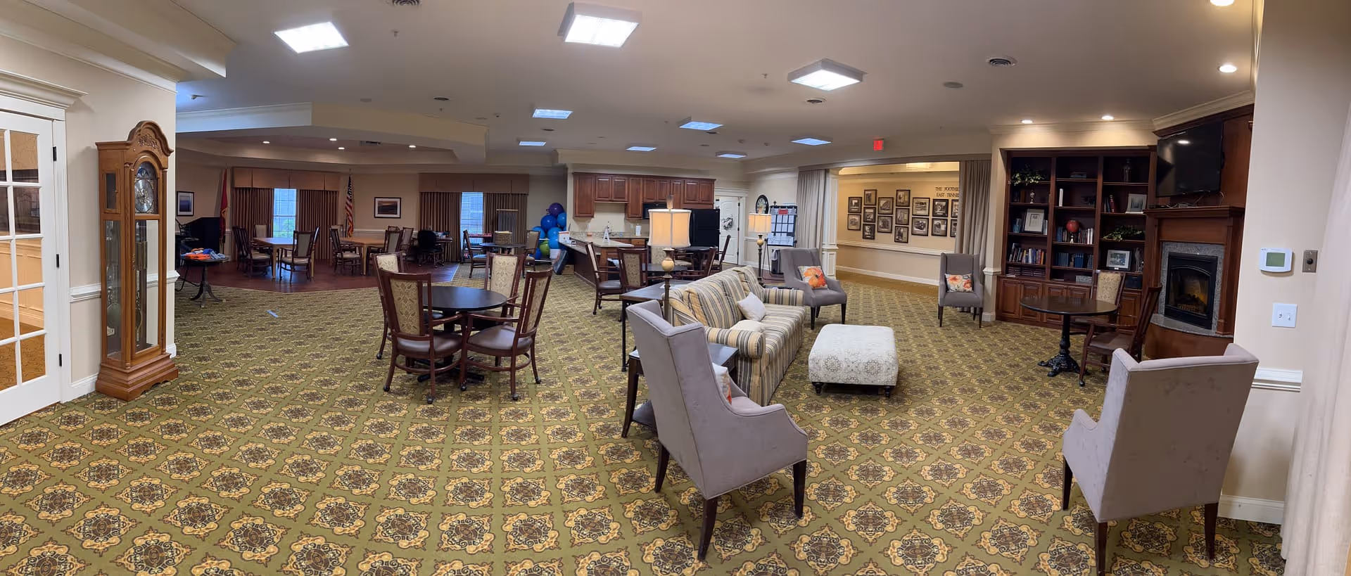 A spacious senior living common area with patterned carpet, multiple seating arrangements including armchairs, a striped sofa, and round tables with chairs. The room features a wooden grandfather clock, a fireplace with a mounted TV above it, built-in bookshelves, and a kitchenette area with wooden cabinets. The walls are decorated with framed pictures and there are ceiling lights providing illumination.