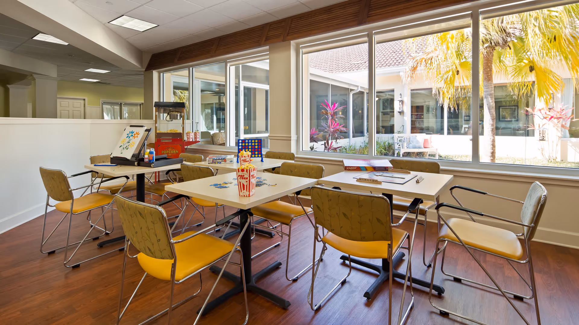 A bright activity room with several tables and chairs arranged for group activities. On the tables are board games, puzzles, and popcorn containers. Large windows provide a view of an outdoor courtyard with palm trees and plants.