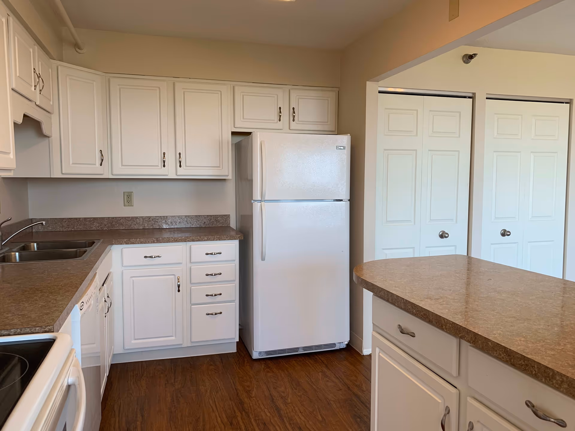 A clean kitchen with white cabinets, a white refrigerator, a double sink, a dishwasher, and a stove. The countertops are brown, and the floor has a wood finish. There are two white closet doors visible in the background.