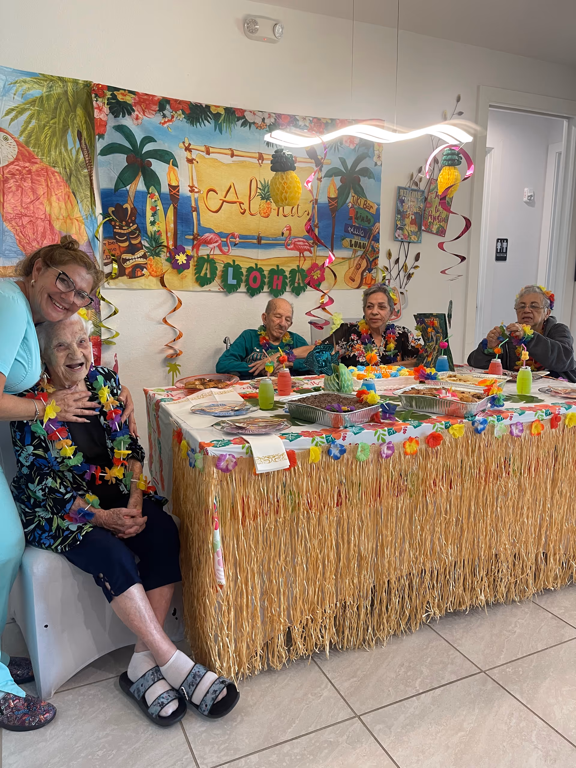 A group of elderly people and a caregiver enjoying a Hawaiian-themed party at a senior living facility. The table is decorated with a grass skirt and colorful flower garlands, and there are tropical decorations on the wall including a banner that says 'Aloha'. The elderly individuals are seated around the table with drinks and food trays in front of them, smiling and engaging in the celebration.