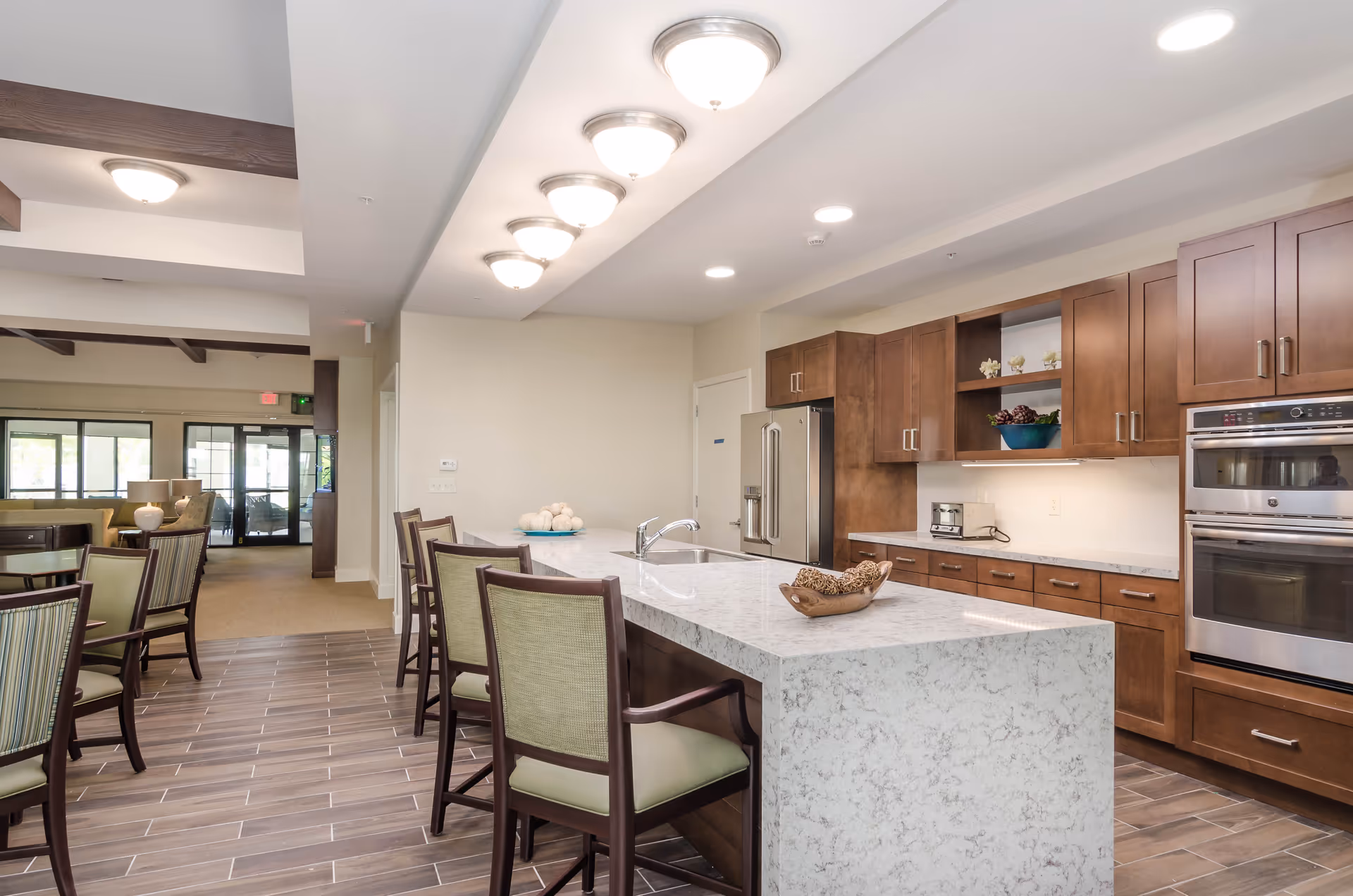 A modern kitchen area in a senior living facility featuring a large marble island with a sink and four chairs. The kitchen has wooden cabinets, a stainless steel refrigerator, double ovens, and a toaster on the counter. The room is well-lit with ceiling lights and has a tiled floor. Adjacent to the kitchen is a dining area with tables and chairs, and a view into a lounge area with large windows and glass doors.