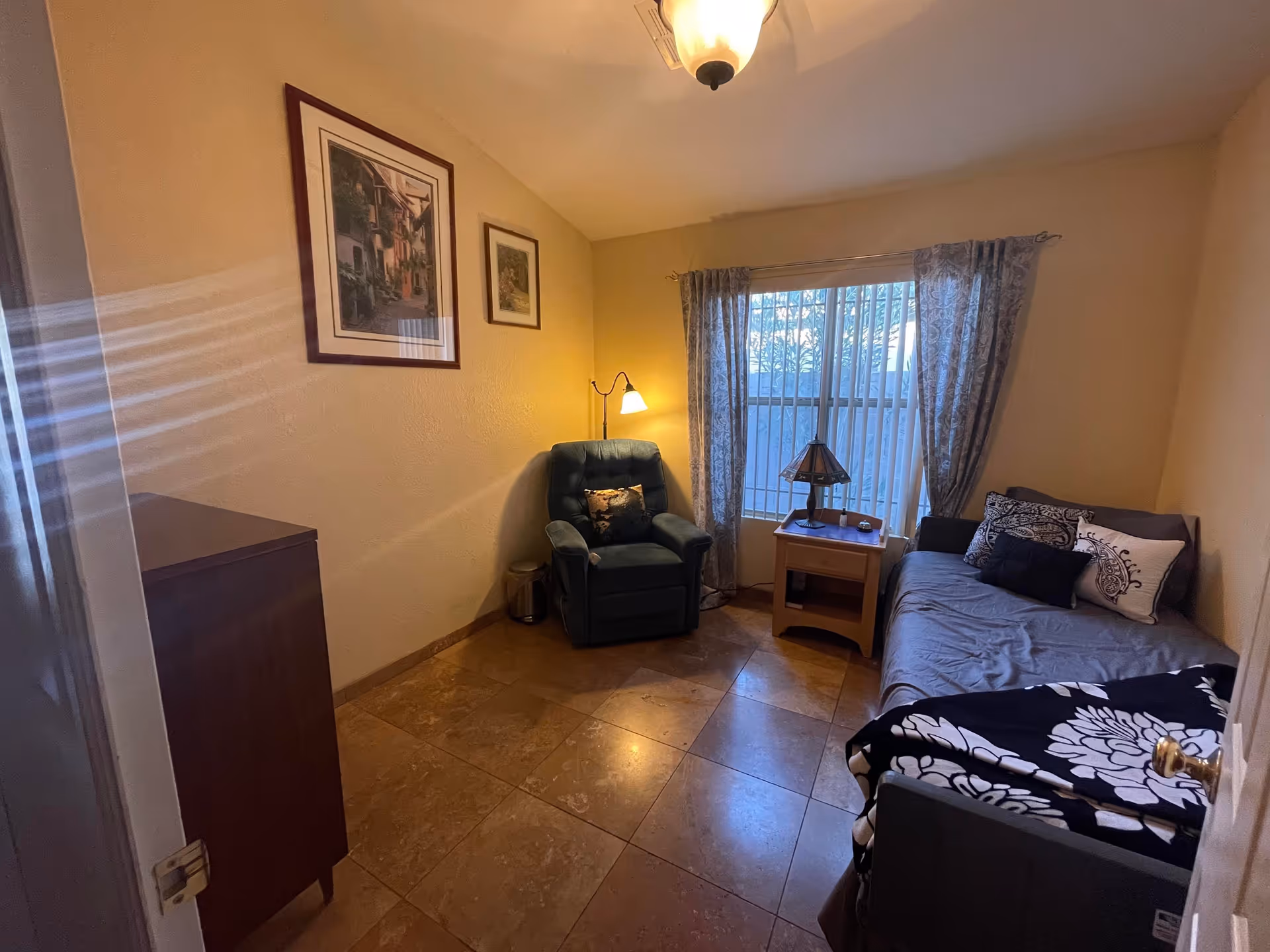 A small, cozy room with a single bed covered in a gray sheet and decorative pillows, a wooden nightstand with a lamp, a green armchair with a floor lamp behind it, two framed pictures on the wall, and a window with patterned curtains letting in natural light.