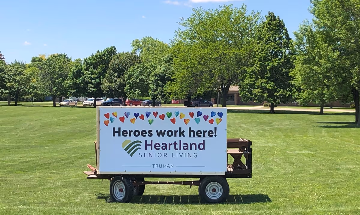 A trailer-mounted sign on a green lawn that reads "Heroes work here! Heartland Senior Living" with trees and parked cars in the background.