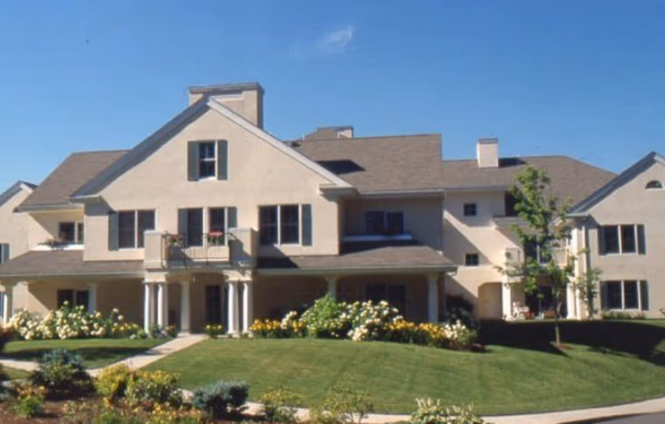 Exterior view of a large, beige senior living facility building with multiple windows, a balcony, and a well-maintained lawn with flower beds and small trees under a clear blue sky.