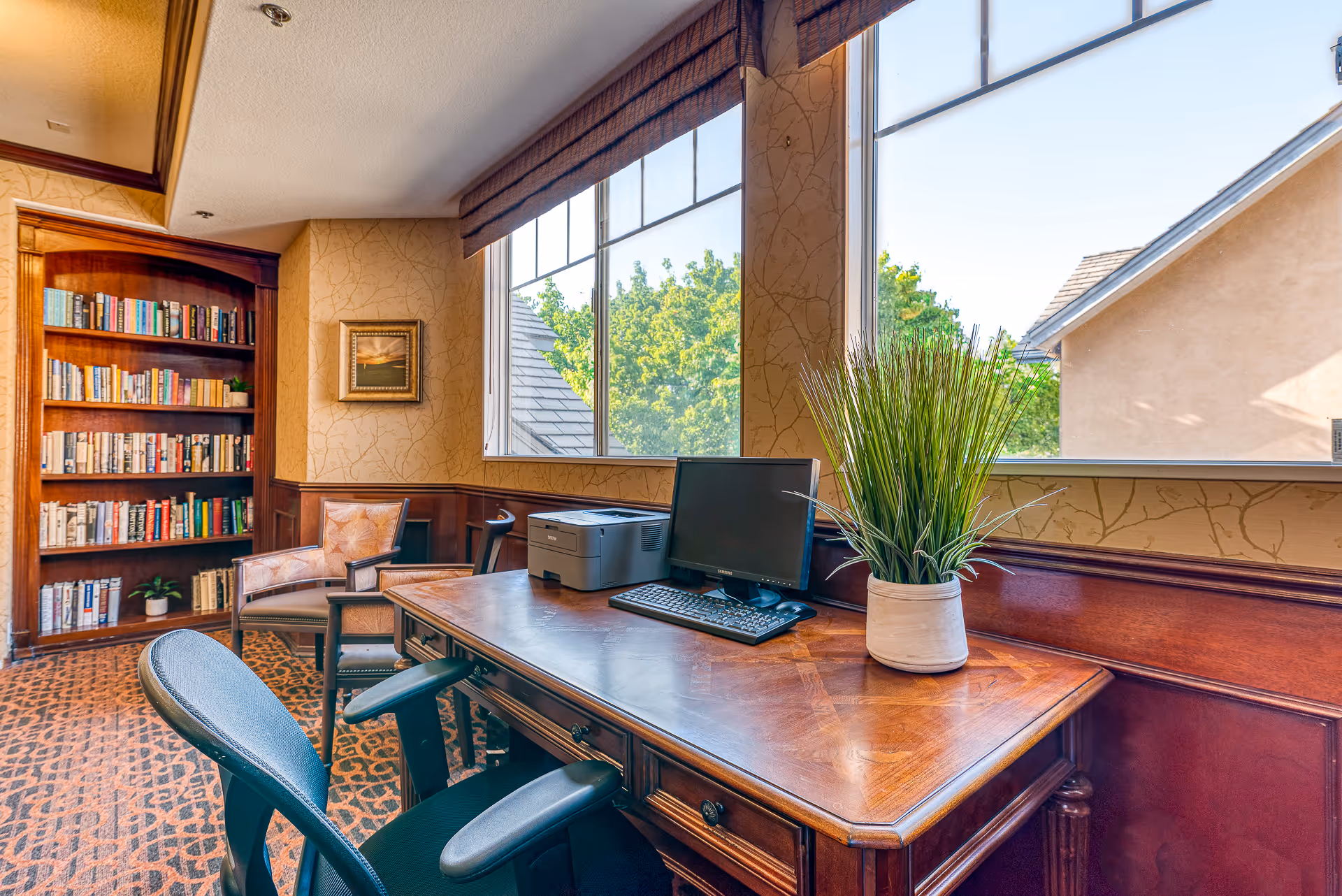 A cozy office or study area in a senior living facility with a wooden desk holding a computer monitor, keyboard, printer, and a potted plant. There are two chairs, a large window with a view of trees and rooftops, and a bookshelf filled with books against the wall.