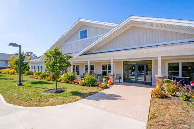 Exterior view of a single-story building with beige siding and a white roof. The entrance features double glass doors with a covered porch supported by white columns. There are landscaped flower beds with shrubs and small trees along the front, and a paved walkway leading to the entrance. The sky is clear and blue.