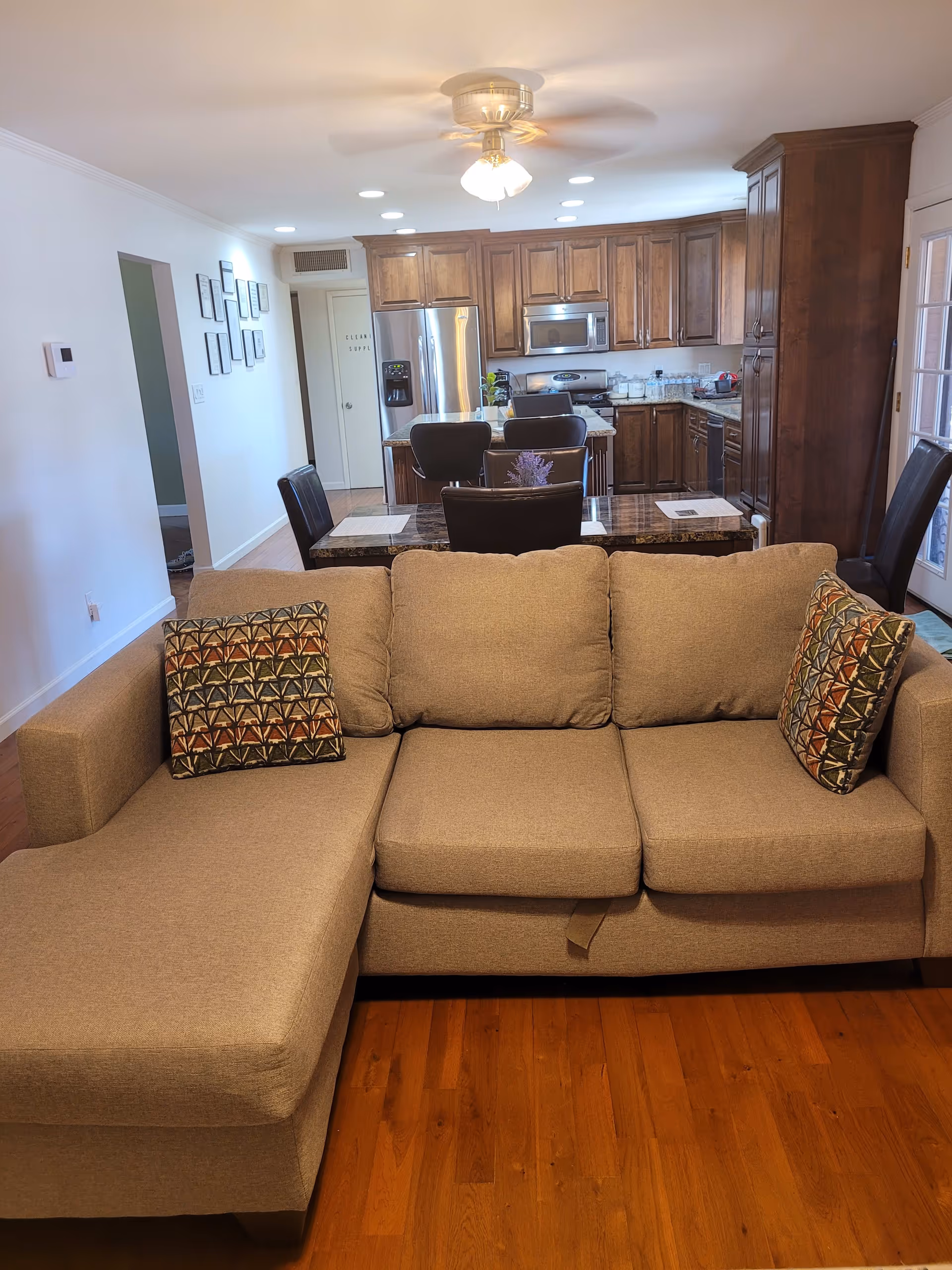 View of a living room area with a beige sectional sofa featuring two patterned pillows. Behind the sofa is a dining table with four chairs, and beyond that is a kitchen with wooden cabinets, stainless steel refrigerator, microwave, and stove. The room has hardwood floors, recessed lighting, and a ceiling fan with lights.