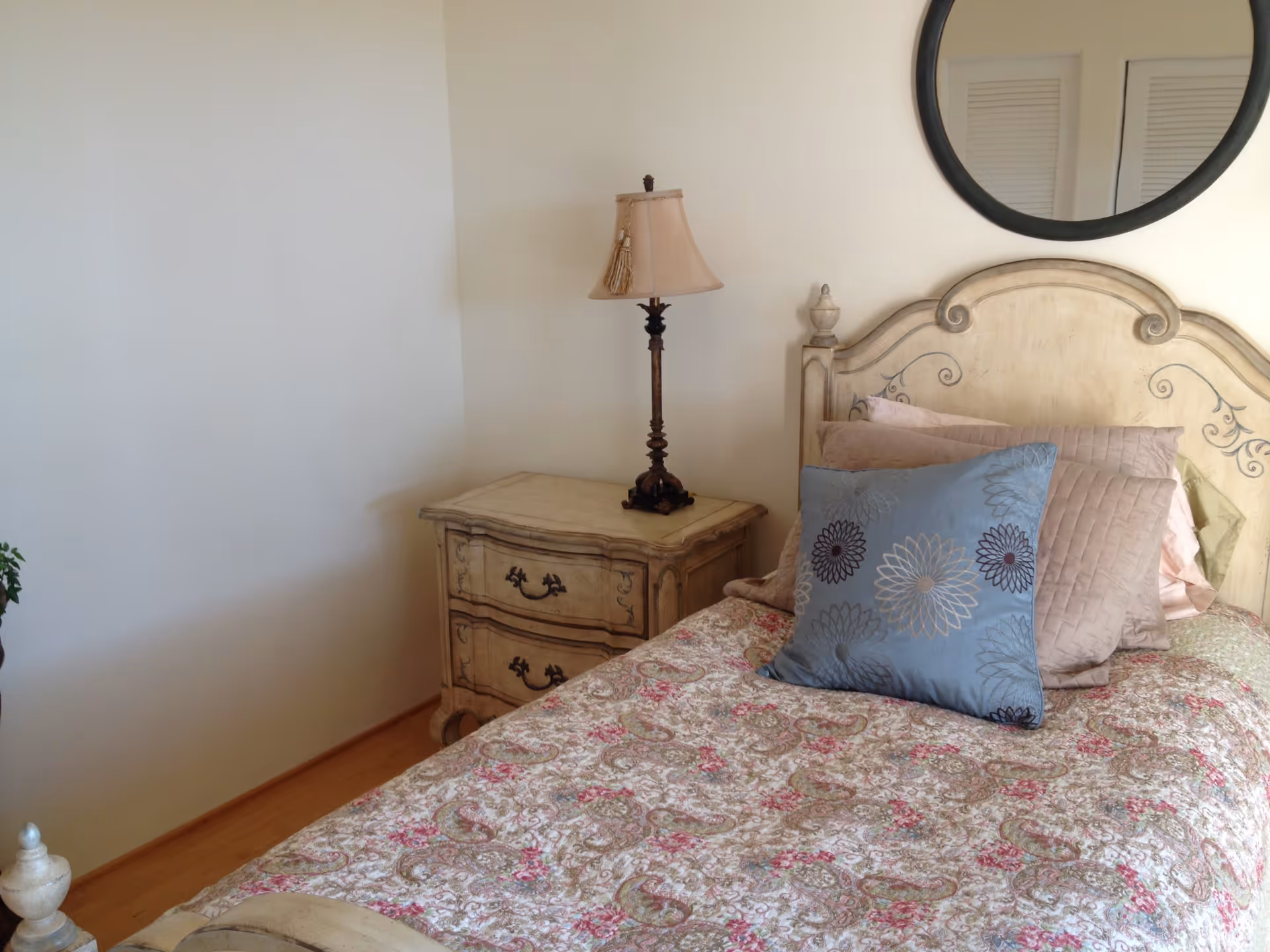 A cozy bedroom featuring a single bed with a decorative floral bedspread and multiple pillows, including a blue one with flower designs. Next to the bed is a vintage-style nightstand with two drawers and an ornate table lamp. A round mirror hangs on the wall above the headboard.