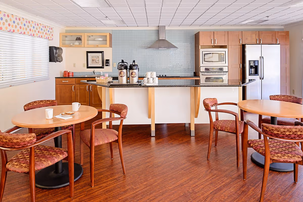 A bright kitchen and dining area with wooden floors, two round tables each surrounded by four patterned chairs, a kitchen island with coffee dispensers and cups, wooden cabinets, a stainless steel refrigerator, microwave, oven, and a range hood against a light blue tiled backsplash.