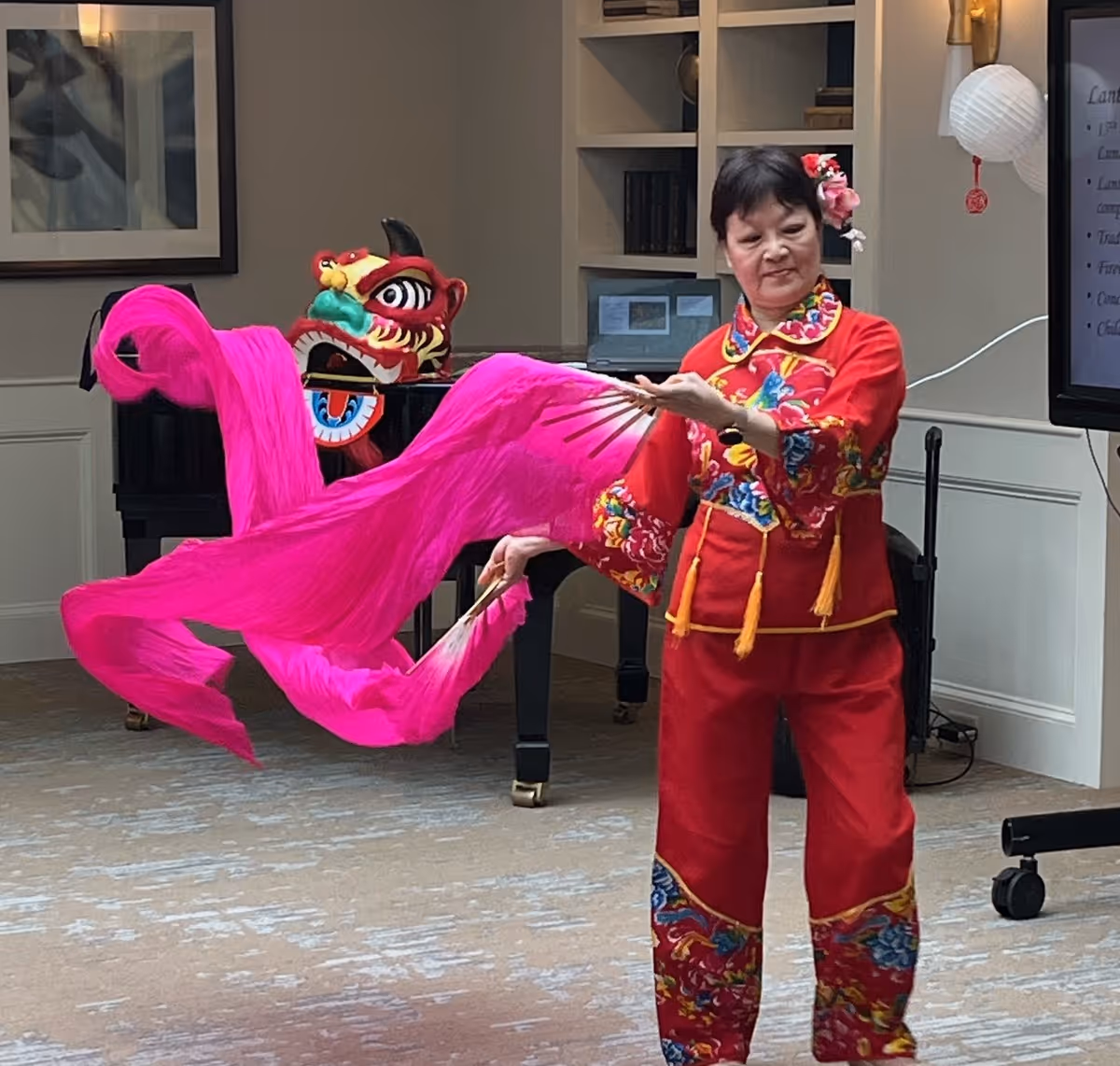 A woman dressed in a traditional red outfit with colorful floral embroidery performs a dance with bright pink flowing fabric fans inside a room. Behind her, there is a piano with a decorative dragon head on it, a bookshelf, and a screen displaying text.