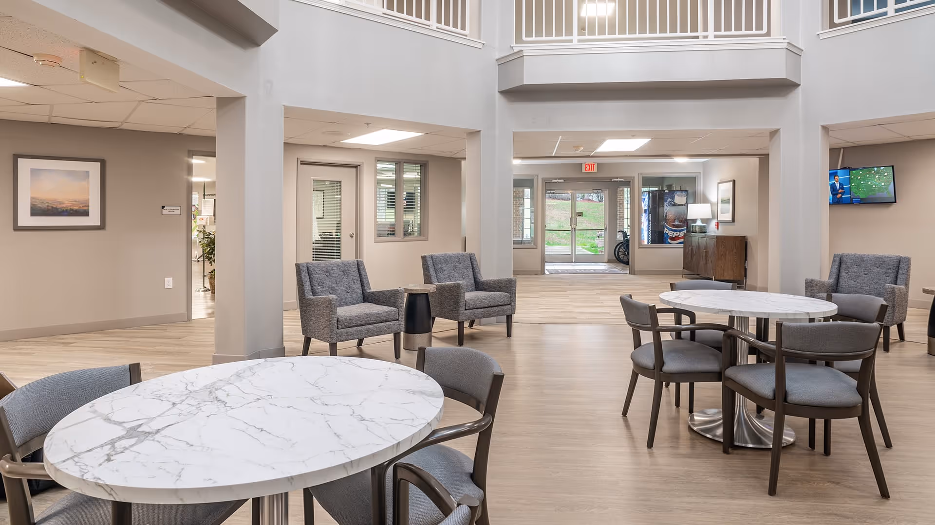 A spacious and well-lit common area in a senior living facility with round marble-top tables surrounded by gray chairs. There are also several gray upholstered armchairs arranged near the walls. The room has light wood flooring, neutral-colored walls, and a high ceiling with a balcony railing visible above. In the background, there is a glass door leading outside and a television mounted on the wall.