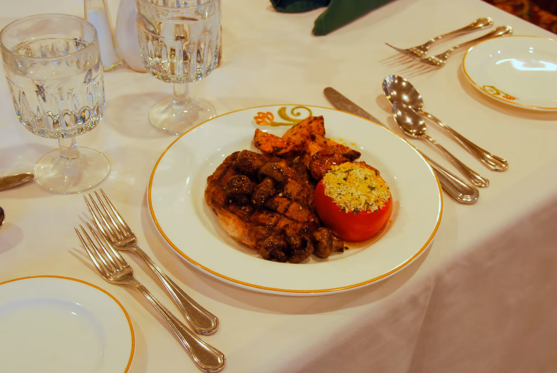 A formal dining table setting with a plate of food including a grilled steak topped with mushrooms, roasted potato wedges, and a stuffed tomato. The table is covered with a white tablecloth and set with crystal water glasses, silver forks, spoons, and knives, along with an empty plate with a gold rim.