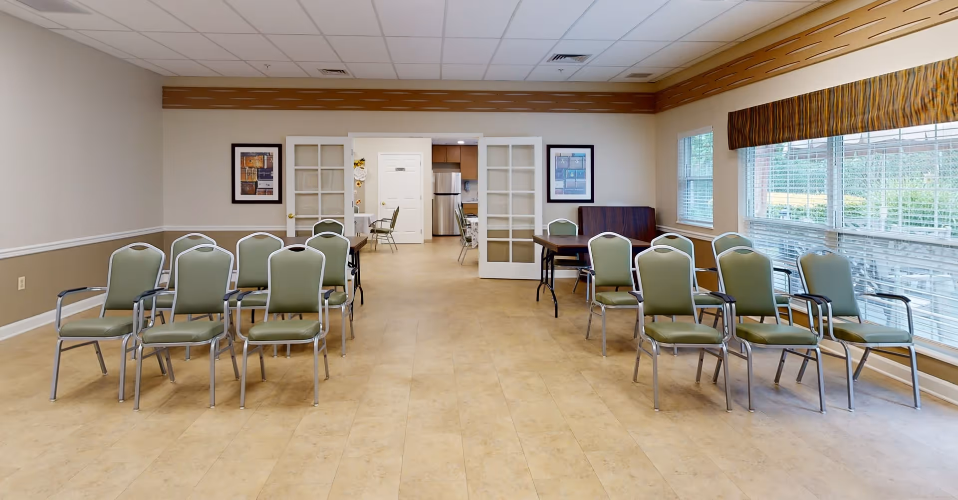 A spacious room with beige walls and tiled floor, featuring two groups of green cushioned chairs arranged in rows facing each other. The room has large windows with blinds on the right side, allowing natural light to enter. At the far end, there are double glass doors leading to another room with a visible refrigerator and additional chairs. Two framed pictures hang on the walls near the doors.
