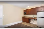 Interior view of a small kitchen area with wooden cabinets, a microwave, a stainless steel refrigerator, and a countertop with a sink. The walls are light beige, and the floor has a combination of carpet and tile.