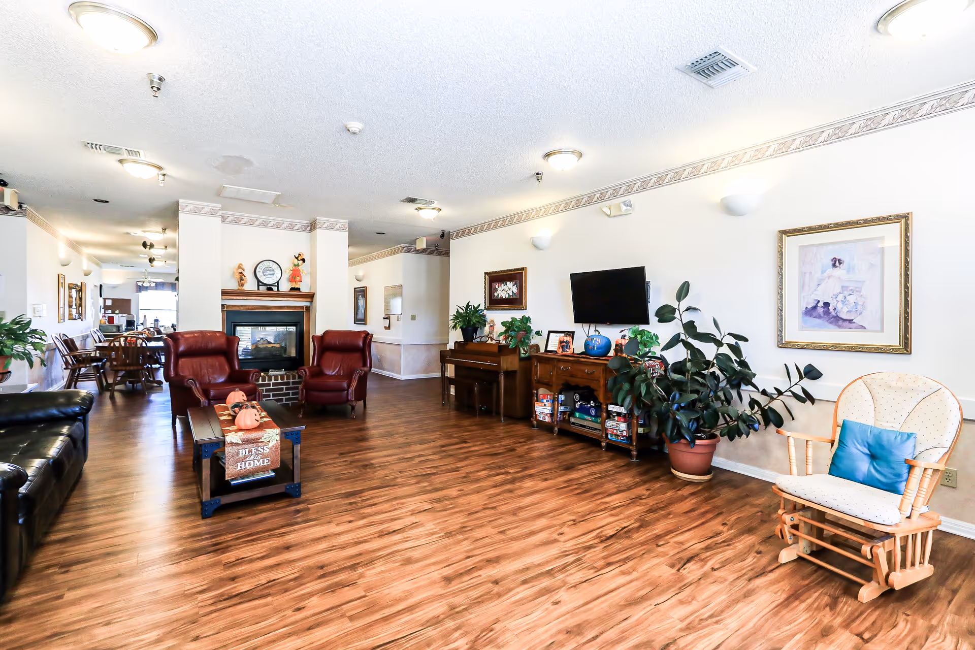 A spacious and well-lit living room area with wooden flooring, featuring two red leather armchairs, a black leather sofa, a wooden coffee table with a decorative sign that says 'Bless this Home', a fireplace with decorative items on the mantel, a piano, a TV mounted on the wall, several potted plants, framed artwork, and a wooden rocking chair with a blue cushion.