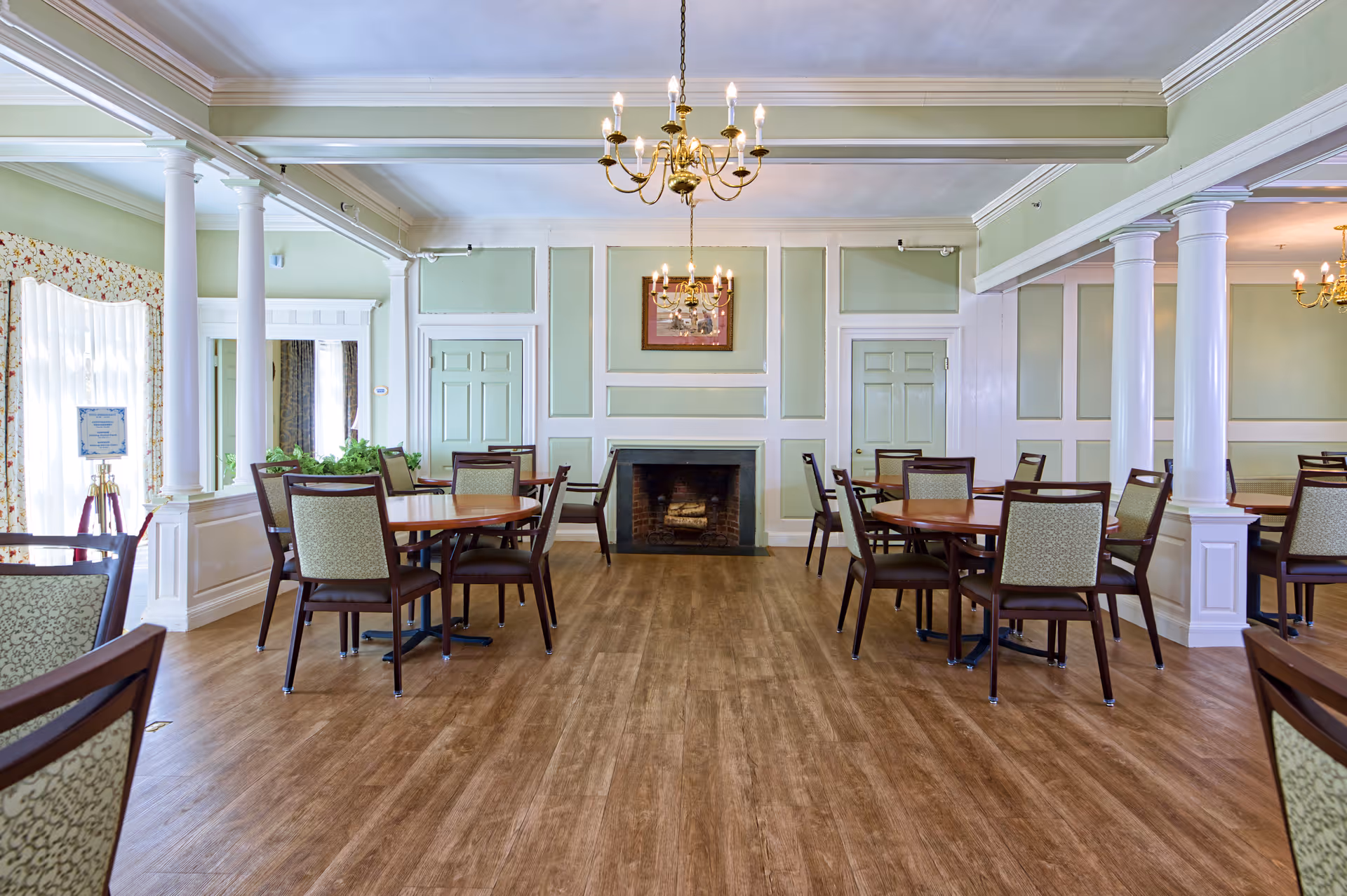 Spacious dining room with round tables and chairs, a central fireplace, chandeliers, and light green paneled walls.