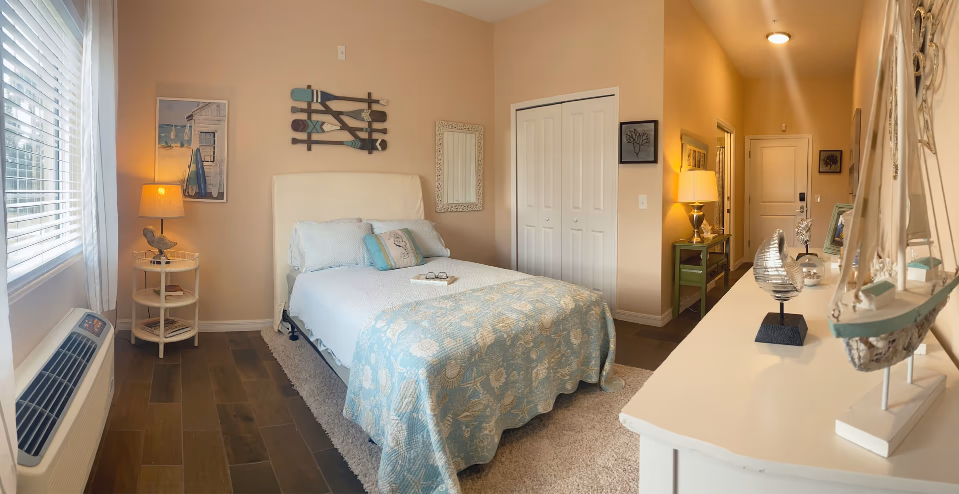 A cozy bedroom with a bed covered in a light blue quilt with seashell patterns. The bed has several pillows and a book with glasses on it. To the left is a window with white blinds and sheer curtains, next to a small round table with a lamp and a bird decoration. On the wall above the bed are decorative wooden oars and a framed mirror. To the right is a white closet door and a hallway with a green console table and a lamp. The room has wooden flooring and soft lighting.