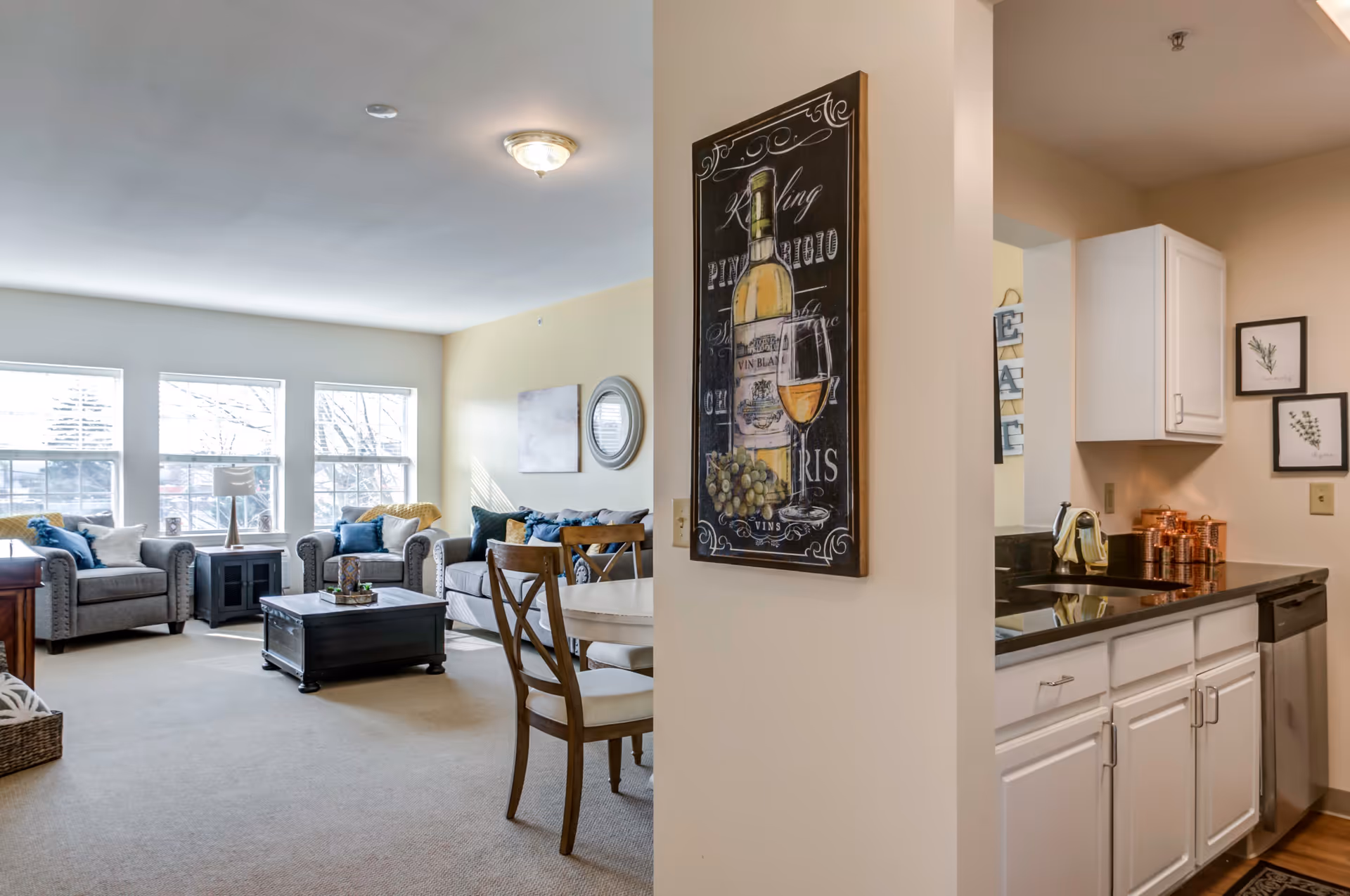 A bright and cozy living area with large windows letting in natural light. The room features two gray armchairs, a gray sofa with blue and yellow cushions, a dark wooden coffee table, and a small side table with a lamp. Adjacent to the living area is a small dining table with wooden chairs. On the right side, there is a kitchen area with white cabinets, a black countertop, a dishwasher, and copper canisters. A decorative wall art featuring a wine bottle and glass hangs on the wall dividing the living and kitchen spaces.