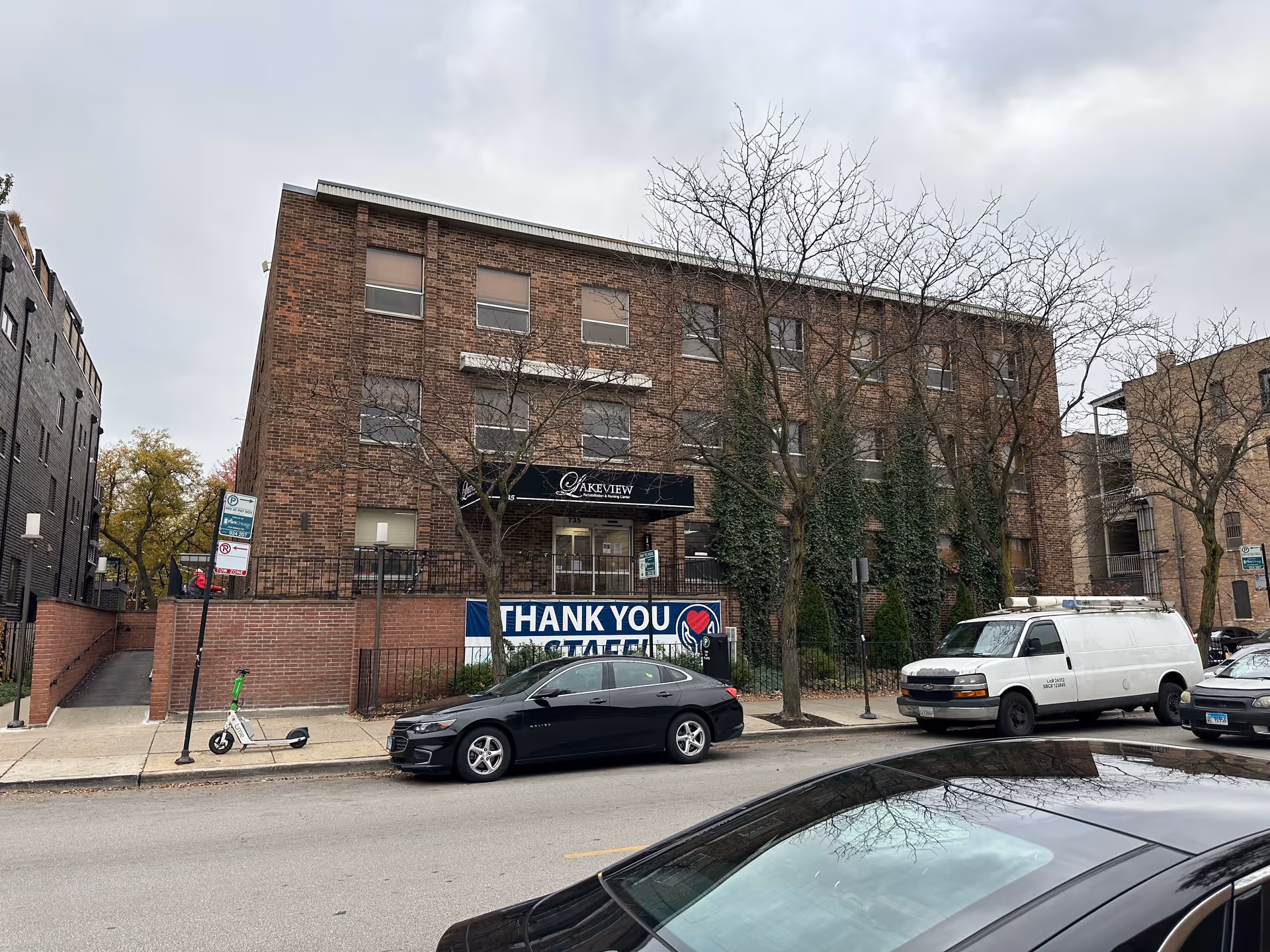 Exterior view of a three-story brick building with a sign that reads 'Lakeview Nursing and Rehabilitation Center' above the entrance. There are leafless trees in front of the building, several parked cars along the street, and a large banner that says 'Thank You Staff'. The sky is overcast.