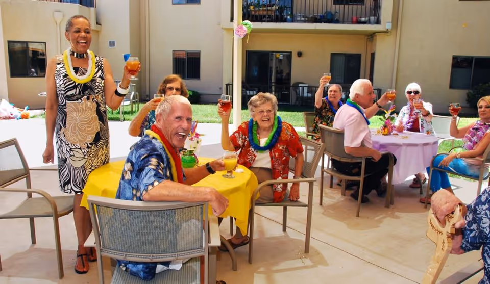A group of elderly people enjoying a cheerful outdoor gathering at a retirement community patio. They are sitting around tables with yellow and white tablecloths, raising their glasses in a toast, wearing colorful leis and smiling.