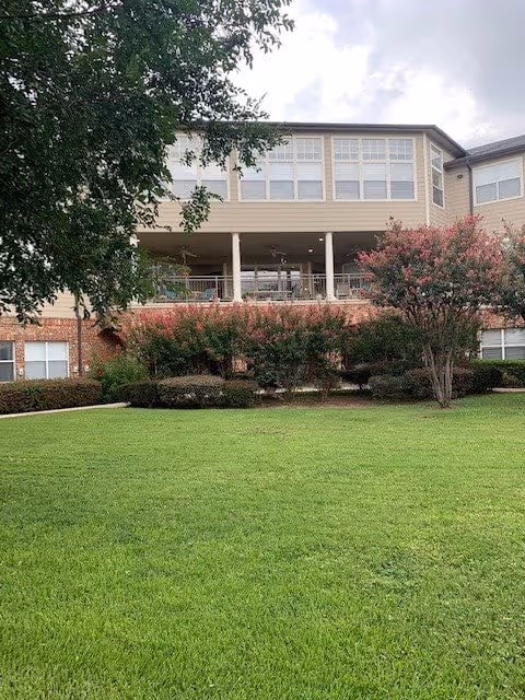 Exterior view of a multi-story senior living facility building with large windows and a covered patio area. The foreground features a well-maintained green lawn, bushes, and trees under a partly cloudy sky.